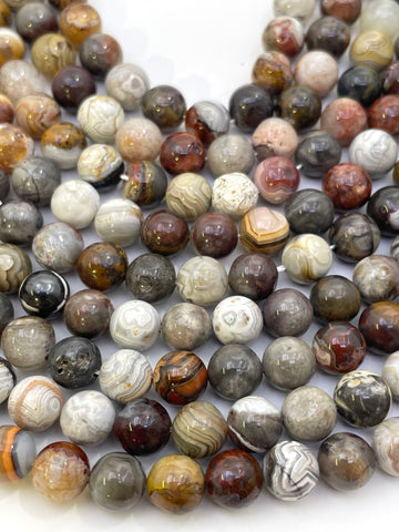 A close-up of many polished round stone mexican crazy agate beads in assorted natural patterns and colors, including gray, brown, cream, black, and reddish tones, arranged on a white background.