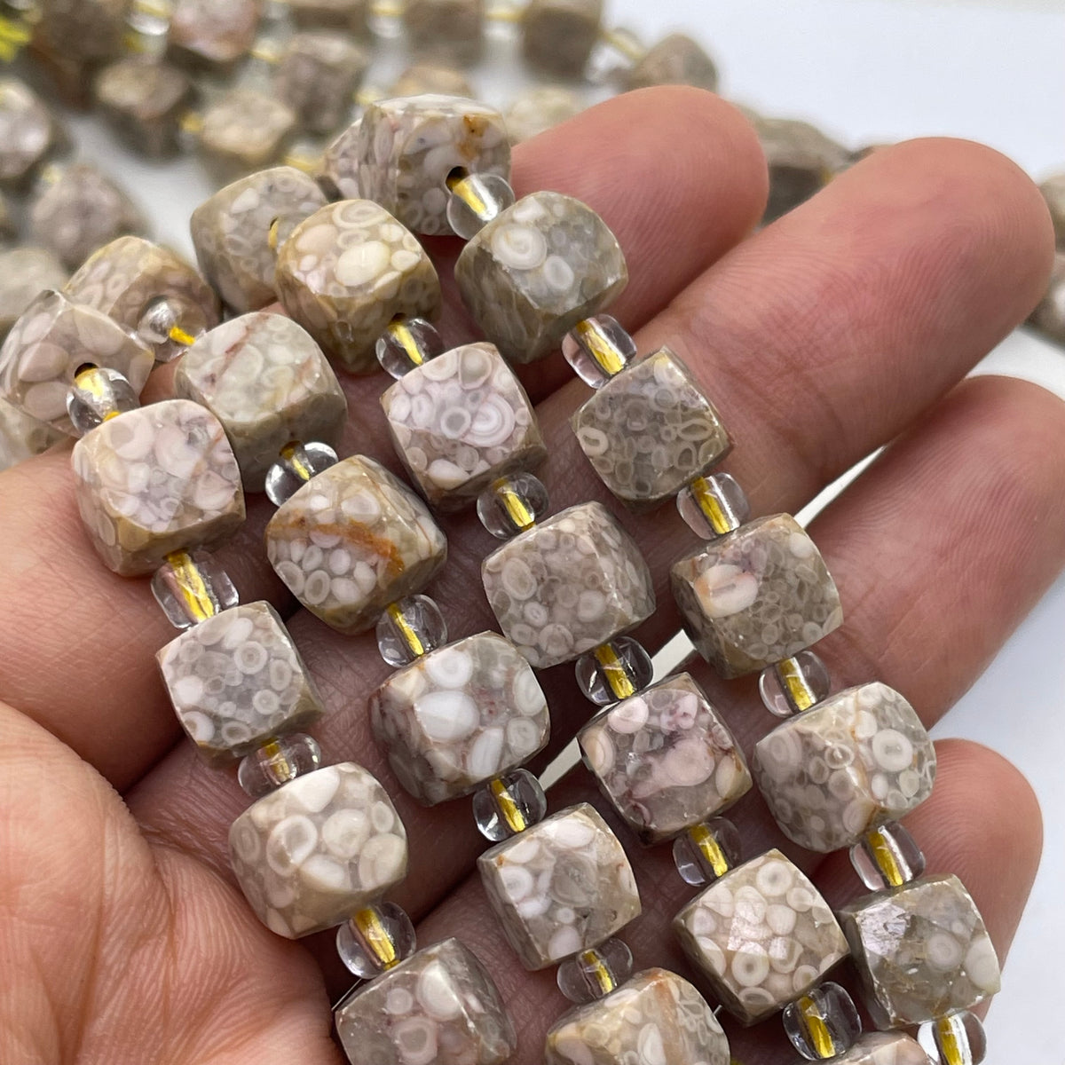 A close-up of a hand holding multiple strands of polished square gemstone beads with a natural banded pattern in beige, white, and gray, separated by clear spacer beads with gold-toned metal links.
