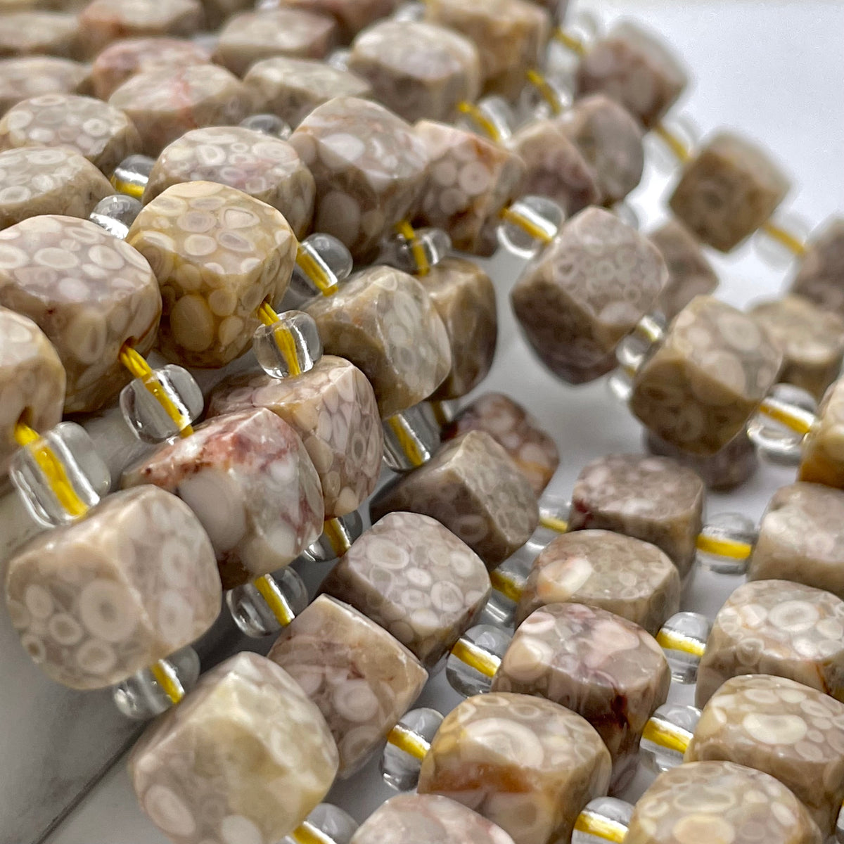 Close-up of polished stone cube beads strung on clear cords with yellow thread, showing a marbled pattern in beige, cream, and brown tones.