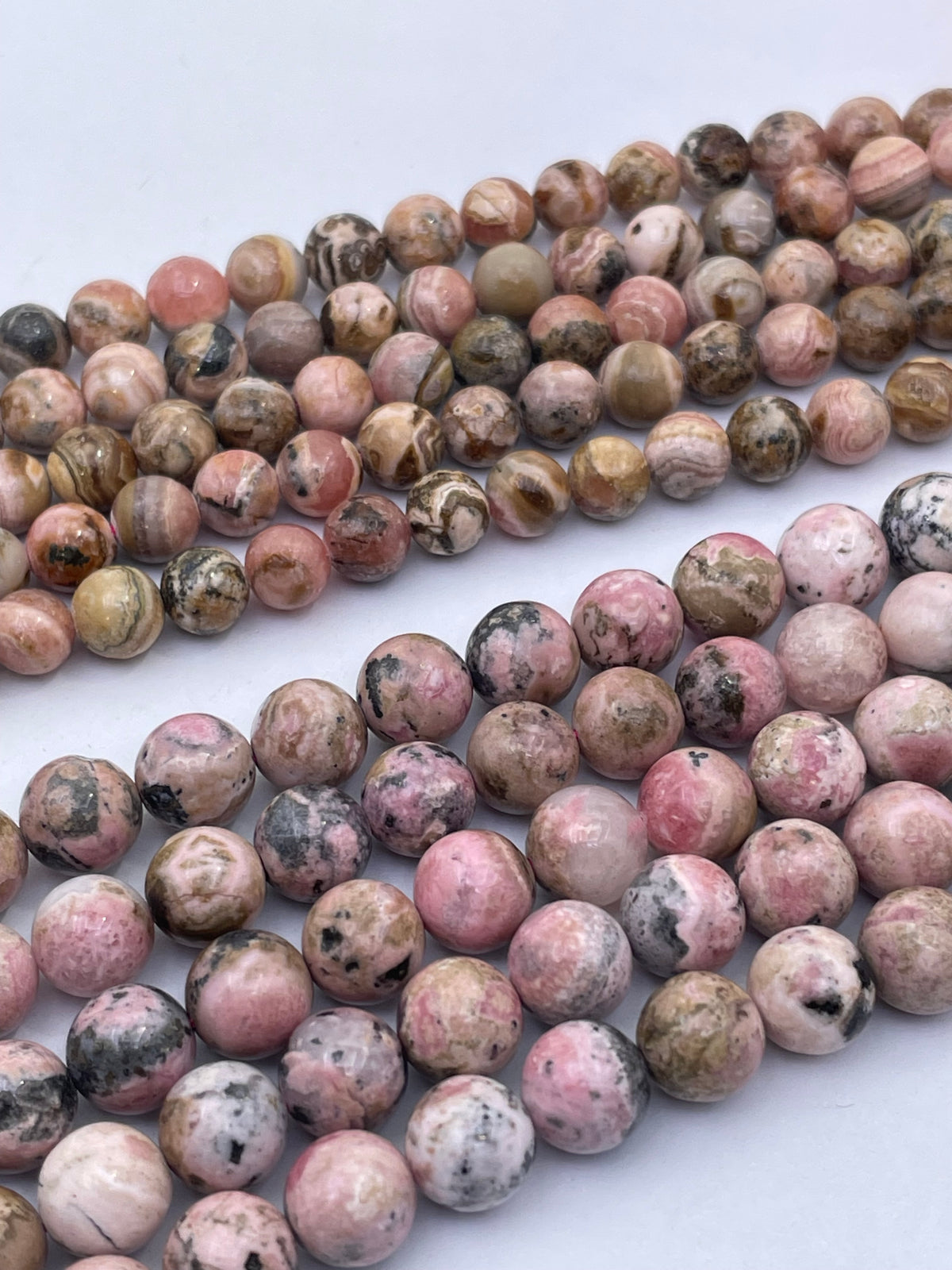 Multiple strands of round polished rhodochrosite gemstone beads in pink, brown, gray, and white marbled patterns arranged diagonally on a white background.