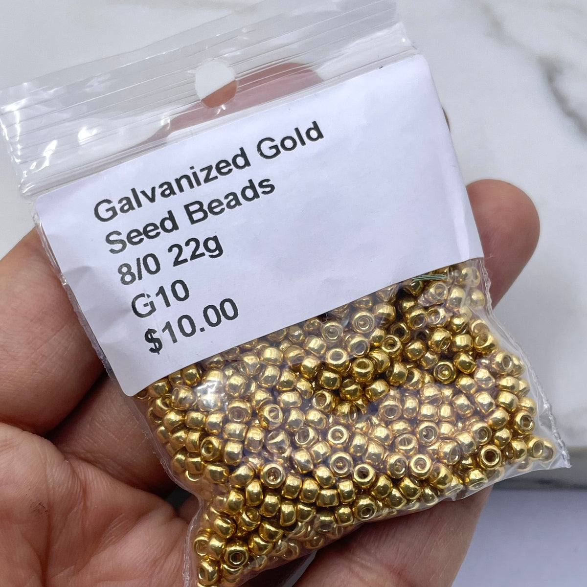 A hand holding a clear plastic bag filled with small galvanized gold seed beads, with a white label reading 'Galvanized Gold Seed Beads 8/0 22g G10 $10.00'.