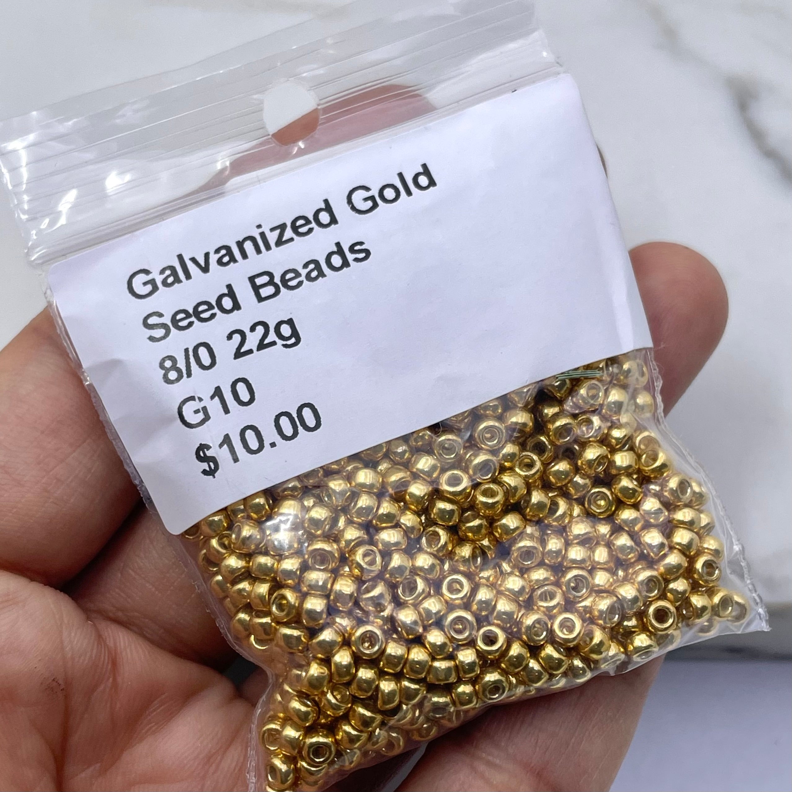 A hand holding a clear plastic bag filled with small galvanized gold seed beads, with a white label reading 'Galvanized Gold Seed Beads 8/0 22g G10 $10.00'.