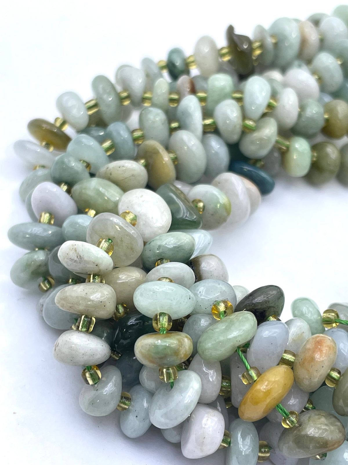 A close-up of a beaded strand made of smooth, polished pebble-shaped burma jade stones in soft sage green, pale gray, white, and light brown tones, separated by small metallic gold spacer beads on a white background.