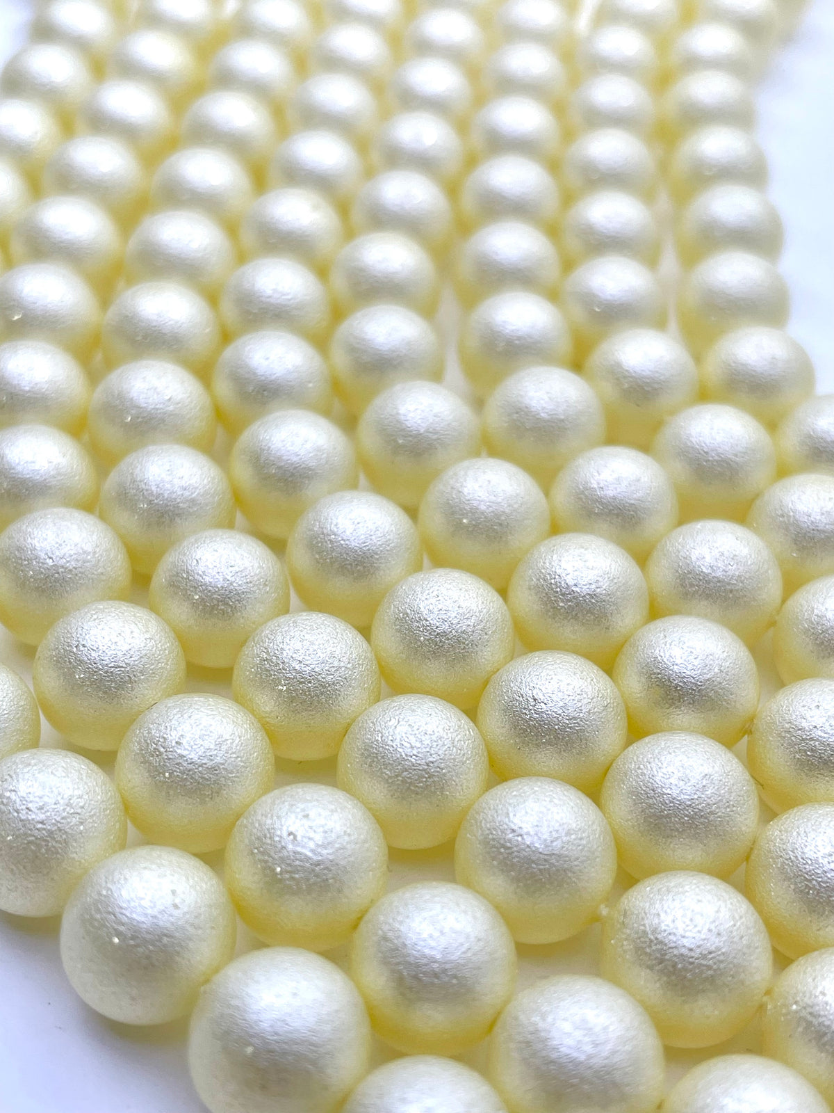A close-up of multiple strands of small, round shell pearl beads in a pale yellow color, arranged diagonally on a white background.