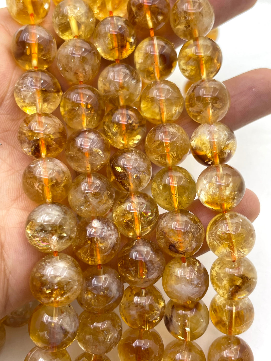 Close-up of round translucent amber-yellow citrine beads strung on cords, showing internal inclusions and a glossy finish against a white background.
