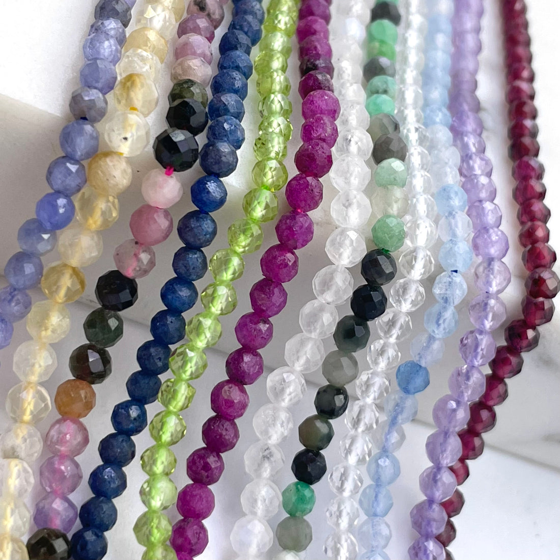 Multiple strands of faceted round gemstone beads in assorted colors, including lavender, yellow, blue, green, pink, purple, gray, and red, arranged vertically on a white background.