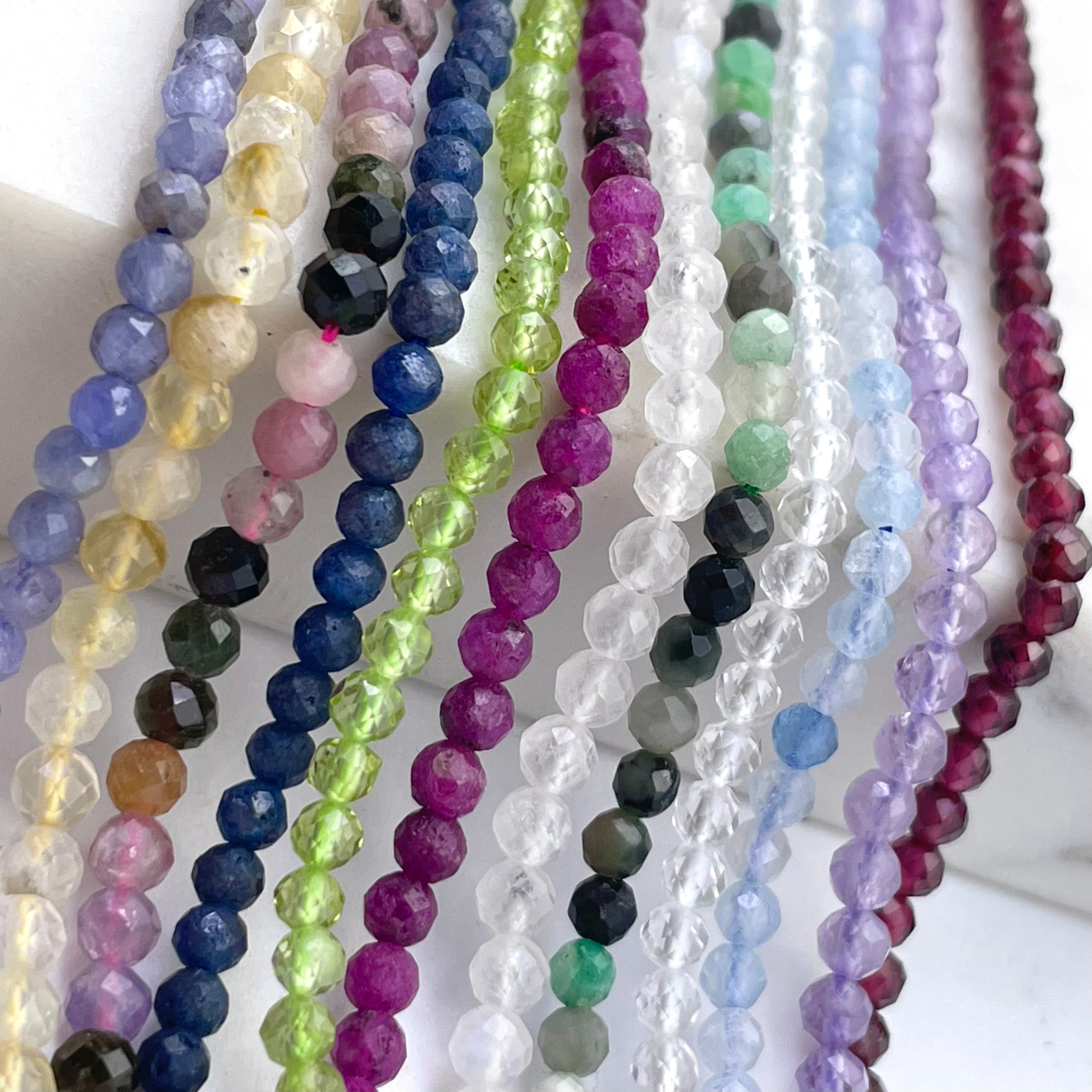 Multiple strands of faceted round gemstone beads in assorted colors, including lavender, yellow, blue, green, pink, purple, gray, and red, arranged vertically on a white background.