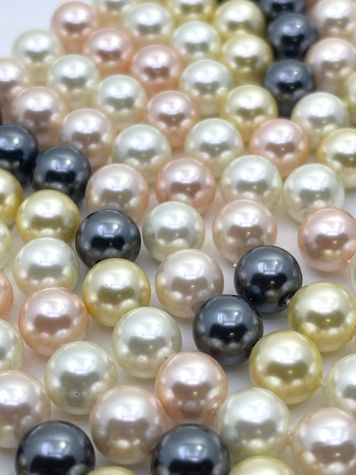 Close-up of a strand of round shell pearls in mixed colors including cream, champagne, peach, and dark charcoal gray, arranged in parallel rows.
