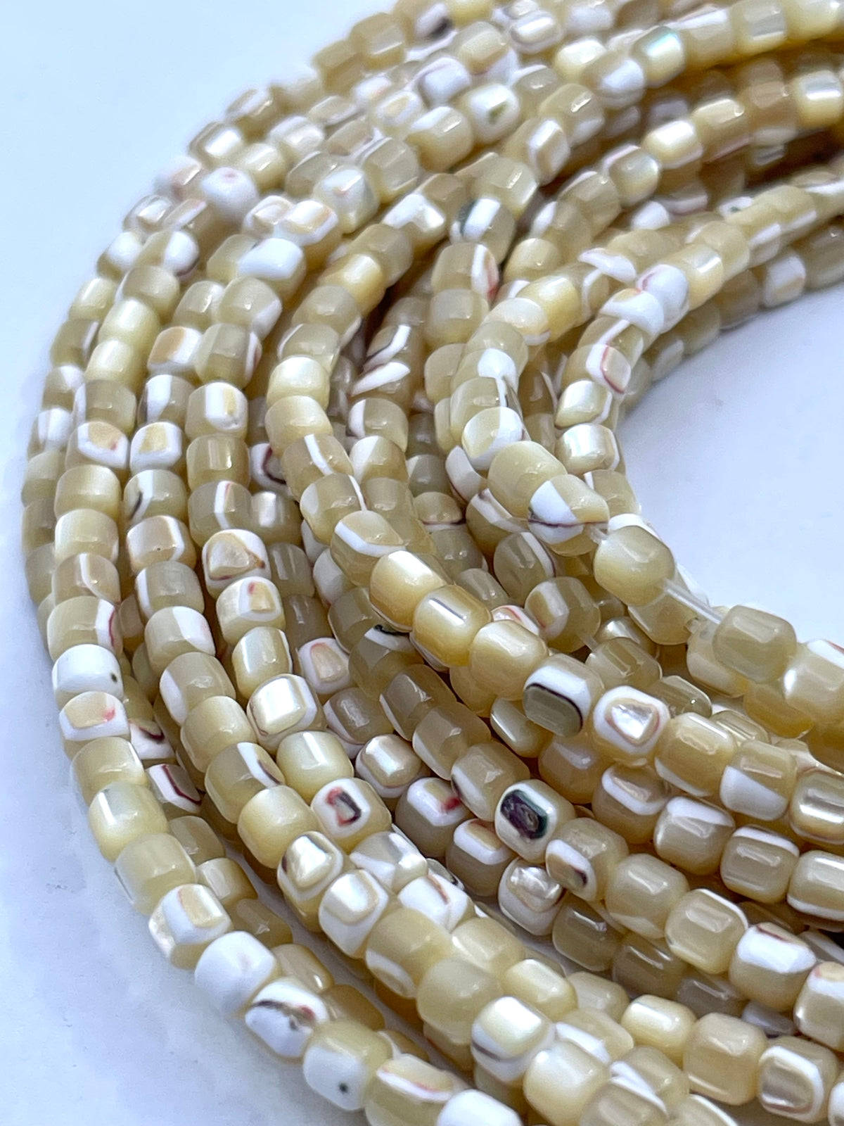 Close-up of multiple strands of small cylindrical mop beads with a glossy finish, featuring a marbled pattern of cream, beige, and white.