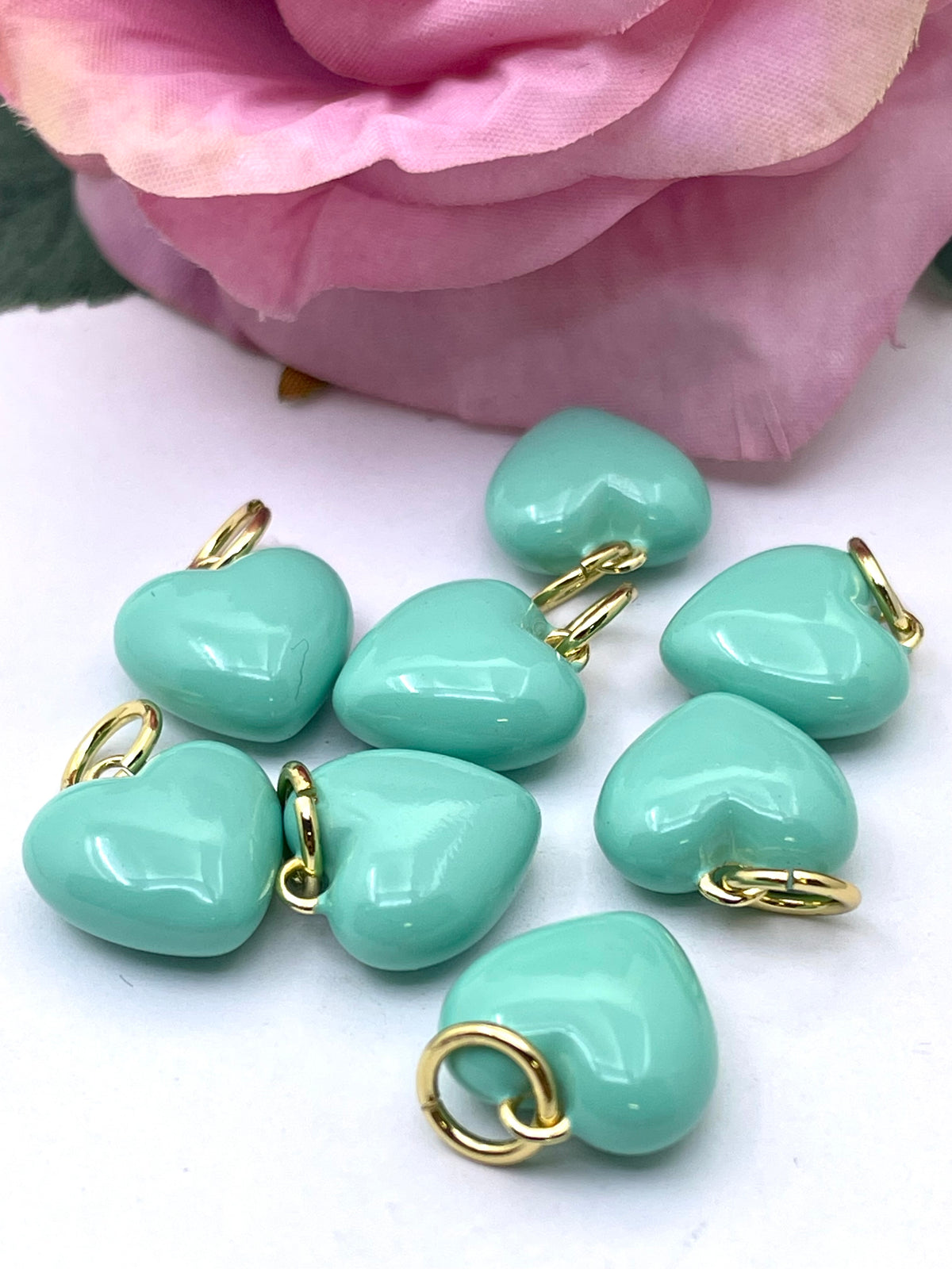Nine glossy mint green heart-shaped charms with gold-colored split rings, arranged on a white surface with a soft pink rose in the background.