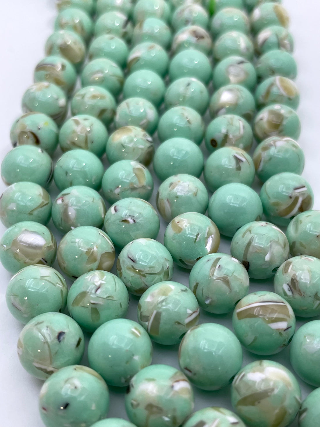 A close-up of multiple strands of round, glossy mop beads in a light mint green color with natural marbled swirls of white, beige, and brown.