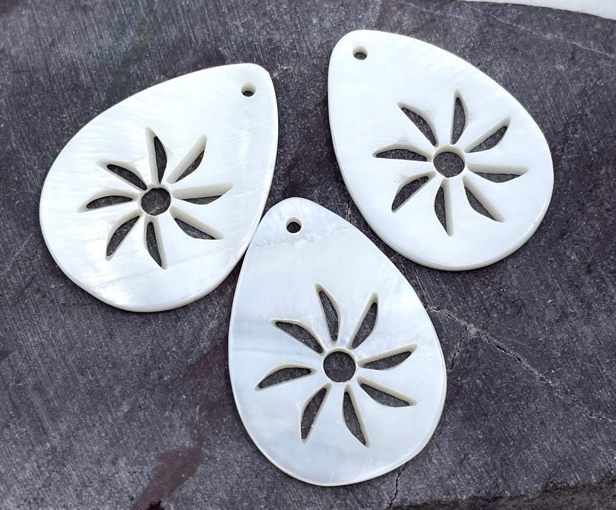 Three white teardrop-shaped shell jewelry charms with a cutout starburst design, displayed on a dark textured slate surface.