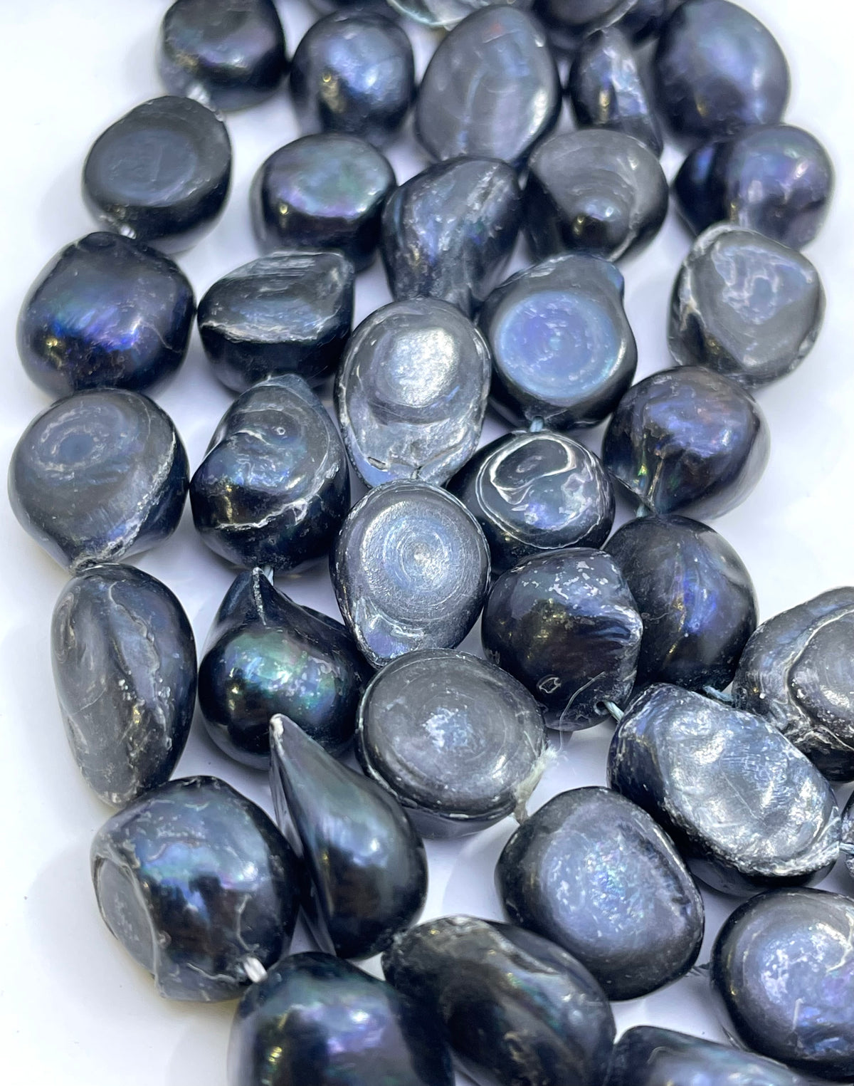 A pile of dark, irregularly shaped polished pearl beads with a pearlescent iridescent finish, showing blue, purple, and gray tones, set against a plain white background.