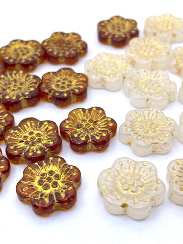 A close-up of flower-shaped beads arranged on a white background, showing two colorways: translucent amber-brown beads with metallic gold foil details, and opaque cream-white beads with matching gold foil details.