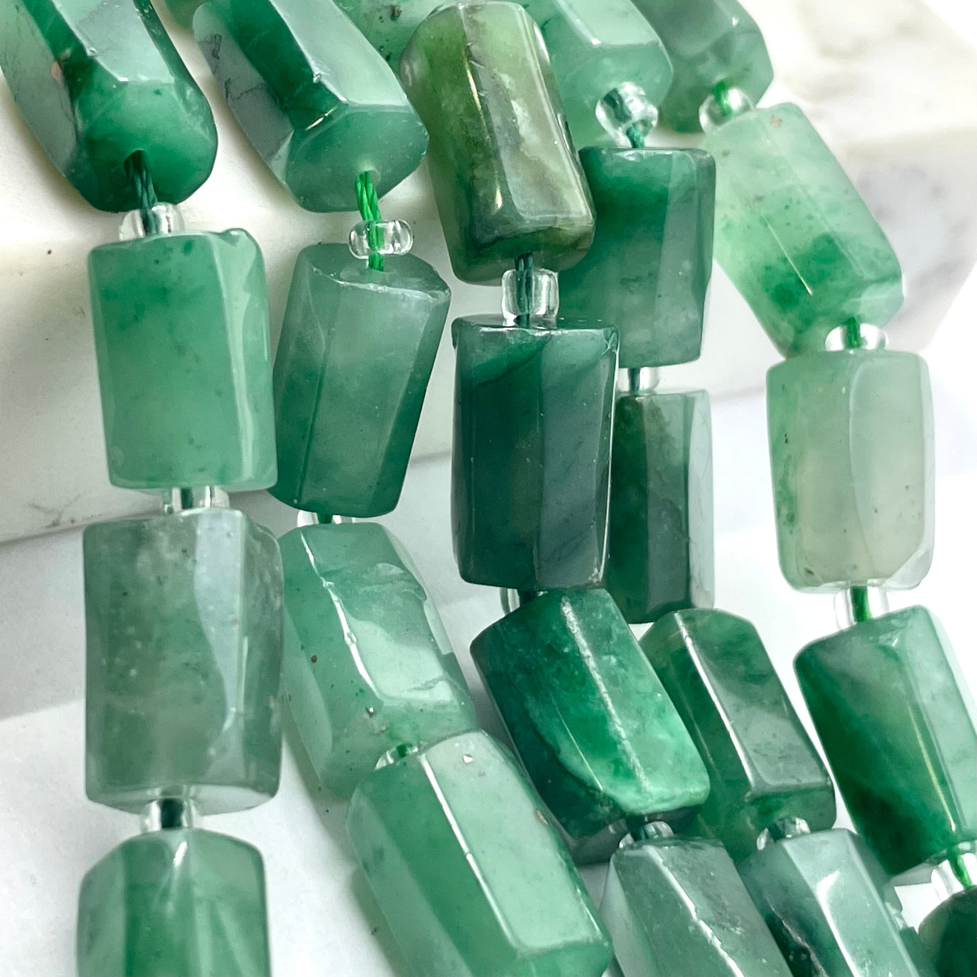 Green jade- rectangular faceted beads strung on silver wire, shown in close-up against a light marble-like background.