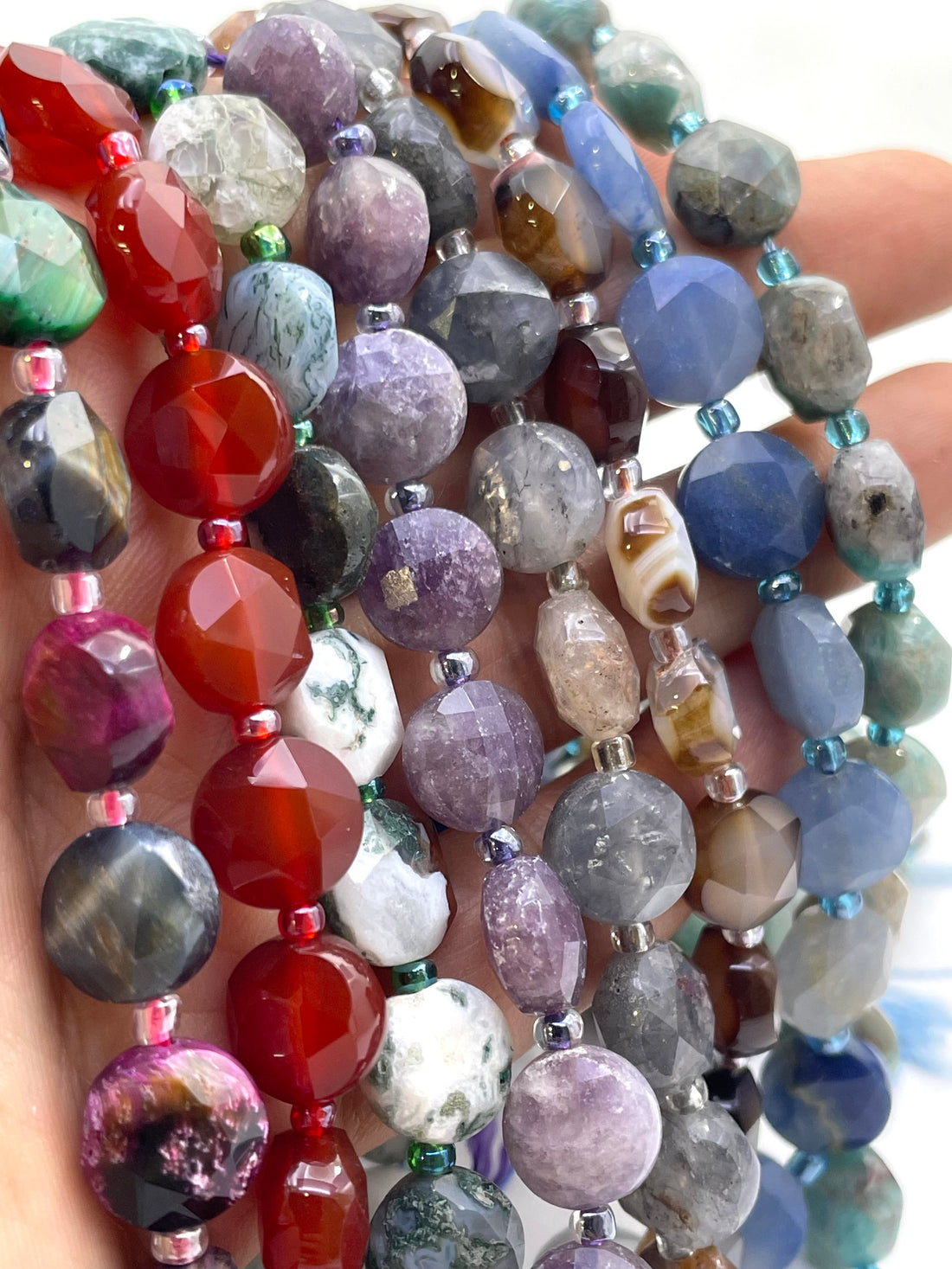 A close-up of multiple strands of faceted gemstone beads held in a hand. The beads come in many colors and patterns including red, purple, blue, gray, green, white, and banded agate designs.