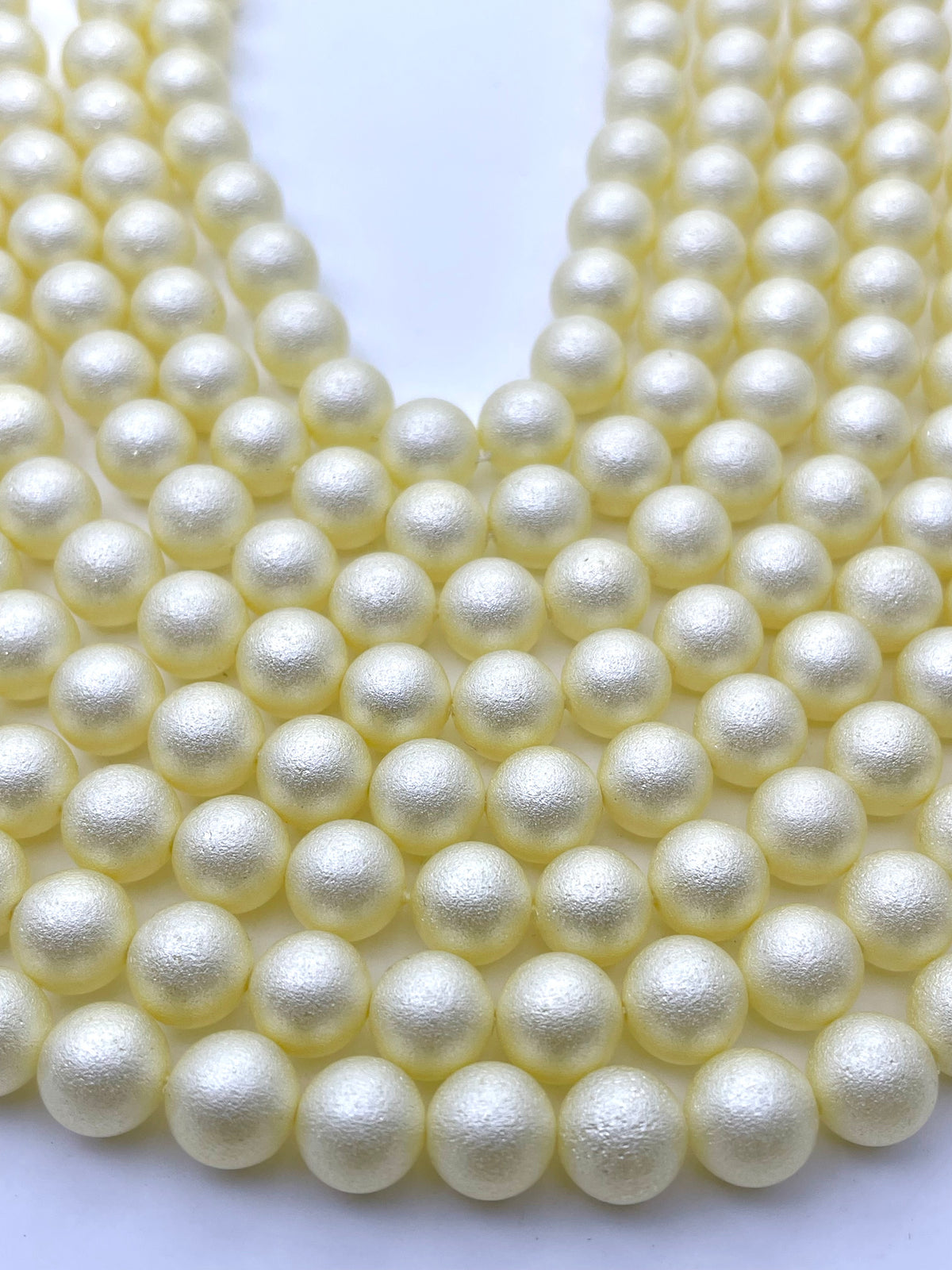 Multiple strands of round shell pearl beads in a warm ivory or pale yellow color, arranged in a V-shape on a plain white background.