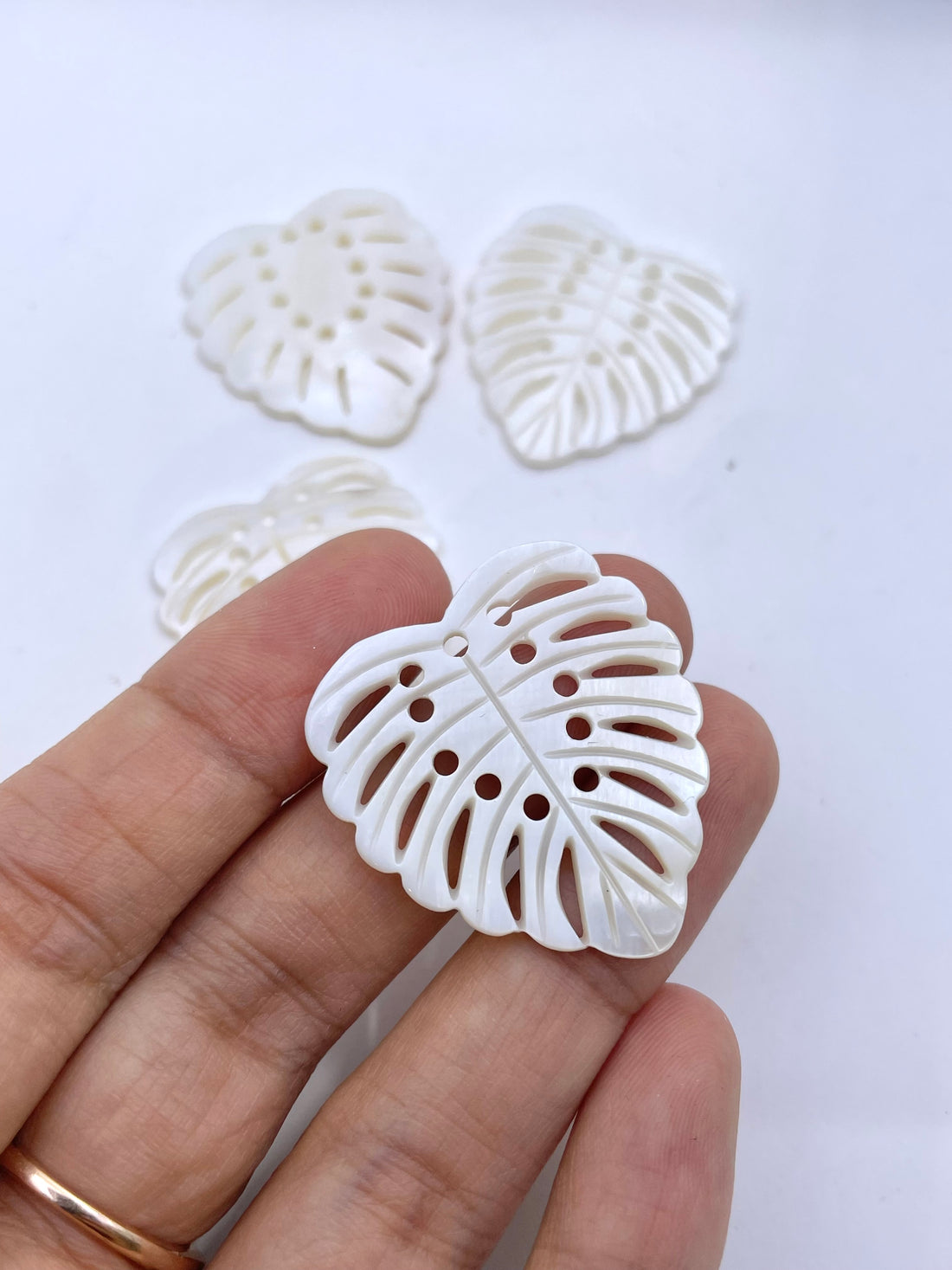 A hand holding a white, leaf-shaped pendant or charm with cutout details, with two more of the same pieces blurred in the background against a white and gray surface.