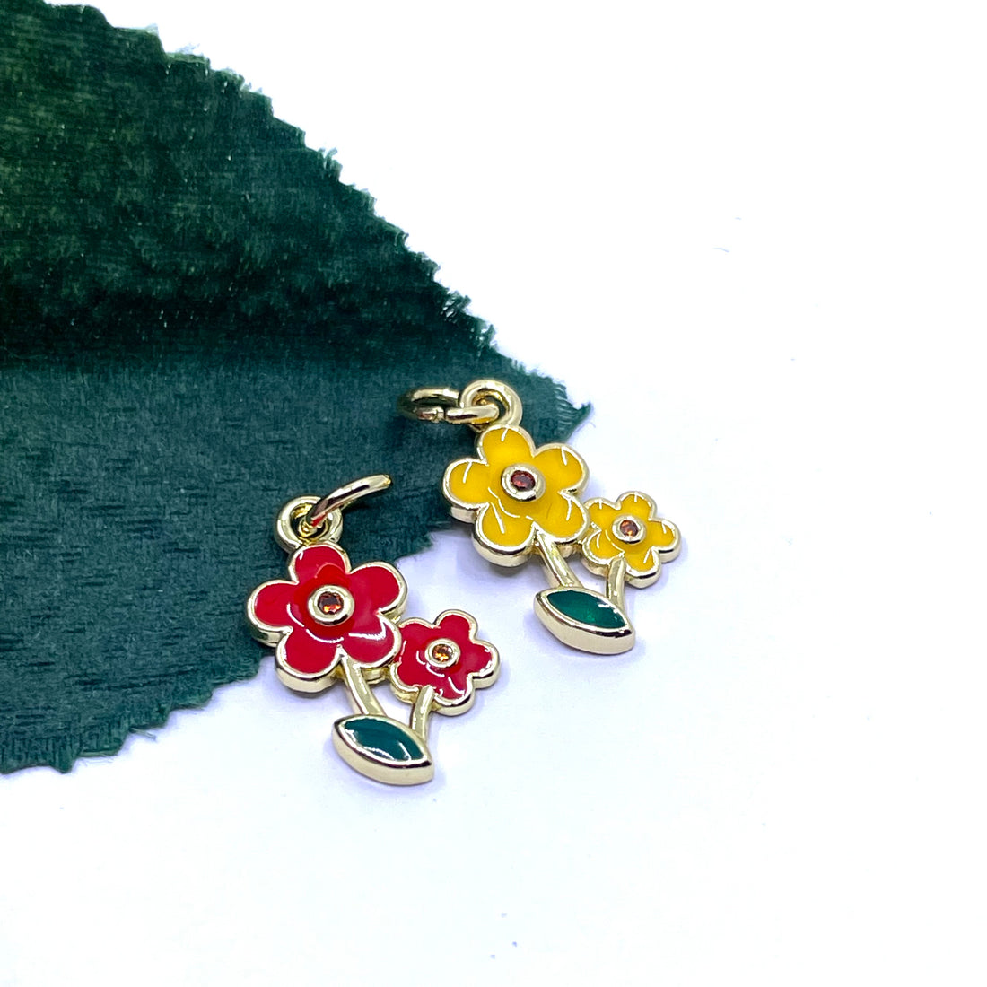 A pair of gold-tone floral dangle charms with red and yellow enamel flowers and green leaves, displayed on a white background with a dark green textured fabric prop.