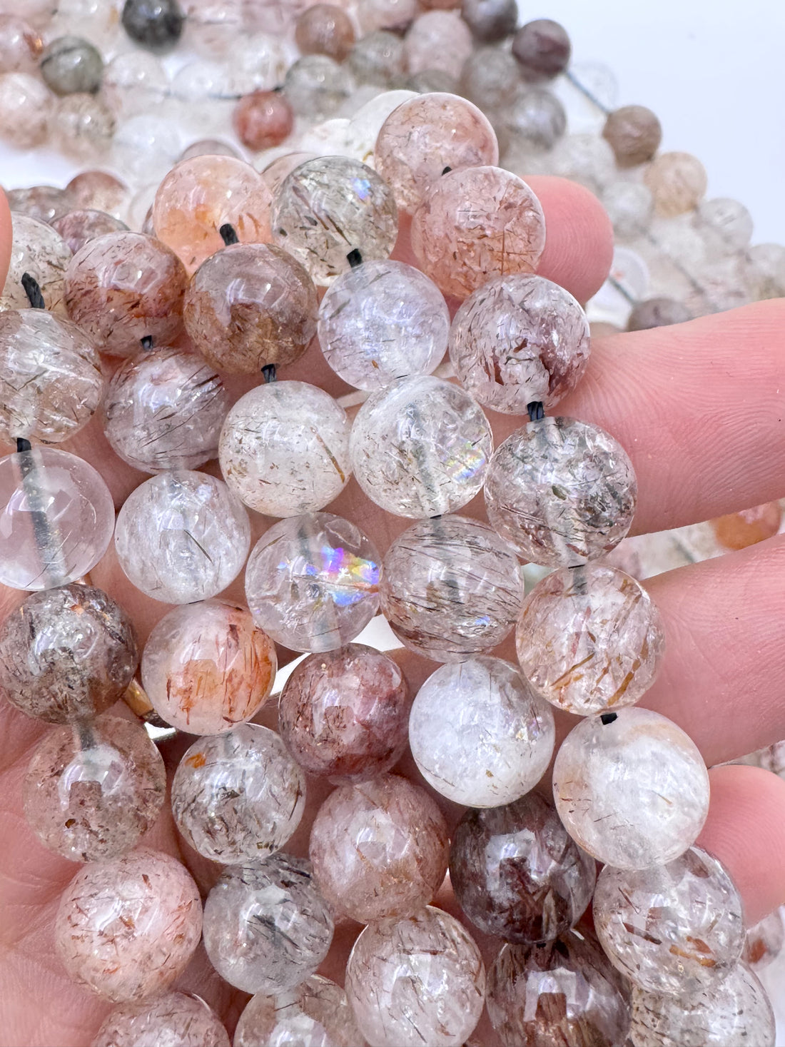 A close-up of a hand holding multiple strands of round black gold super seven quartz bead strands with visible inclusions and some iridescent reflections.
