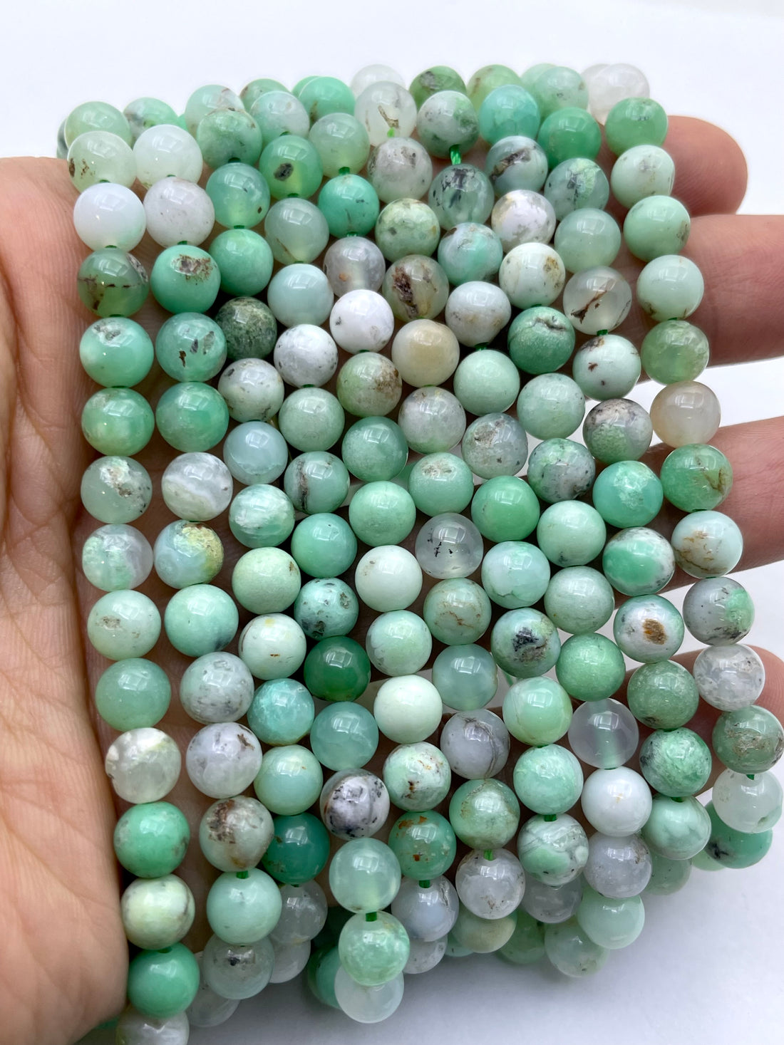 Round natural chrysoprase stone beads in shades of mint green, light aqua, white, and brown, arranged in a curved line on a plain white background.