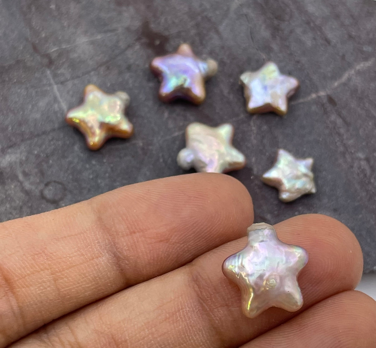 Five small iridescent star-shaped beads or charms resting on a dark textured surface, with a human hand holding one of the stars in the foreground to show scale.