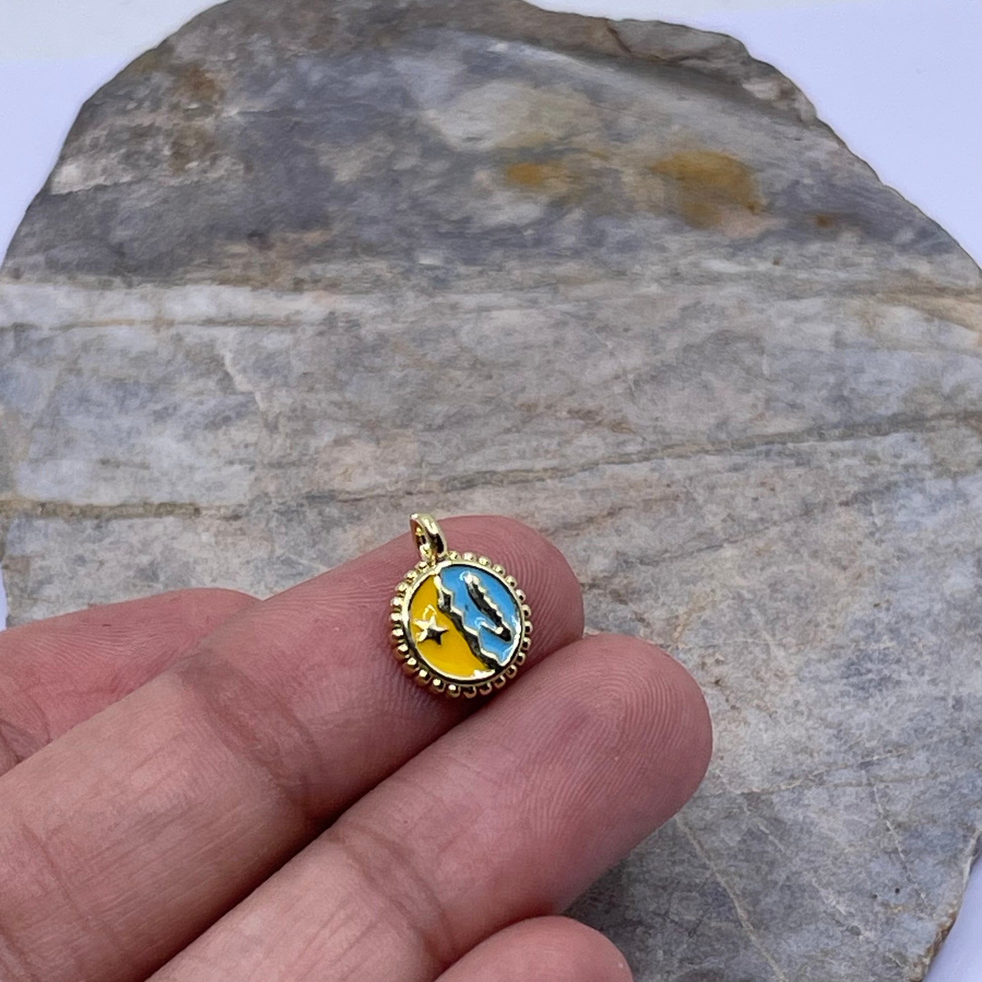 A small round gold pendant with a blue and yellow celestial design, held on a person's fingers against a gray stone background.
