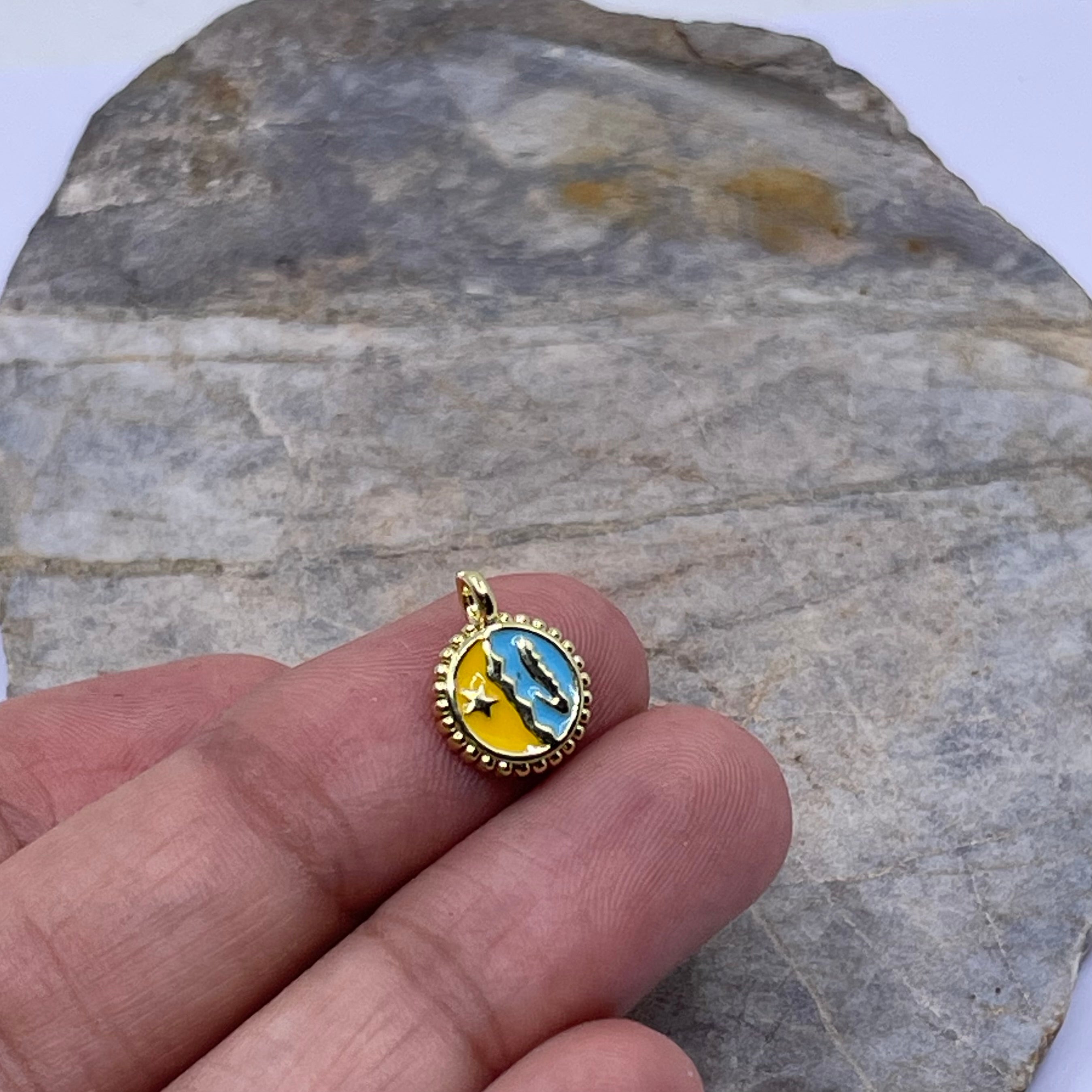 A small round gold pendant with a blue and yellow celestial design, held on a person's fingers against a gray stone background.