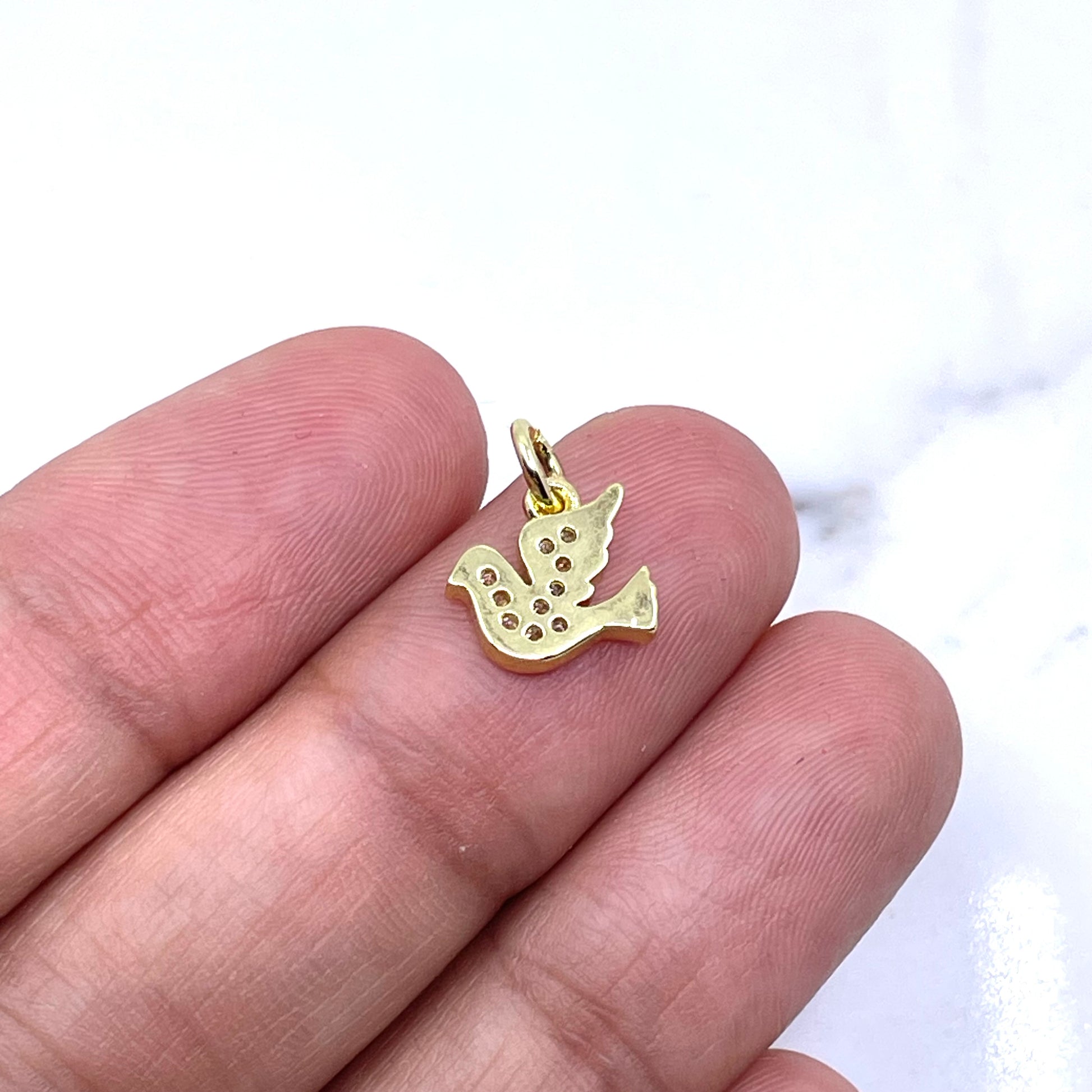 A small gold bird charm with a textured, dotted pattern is held between a person's fingers against a white marble background. (back side of dove charm)