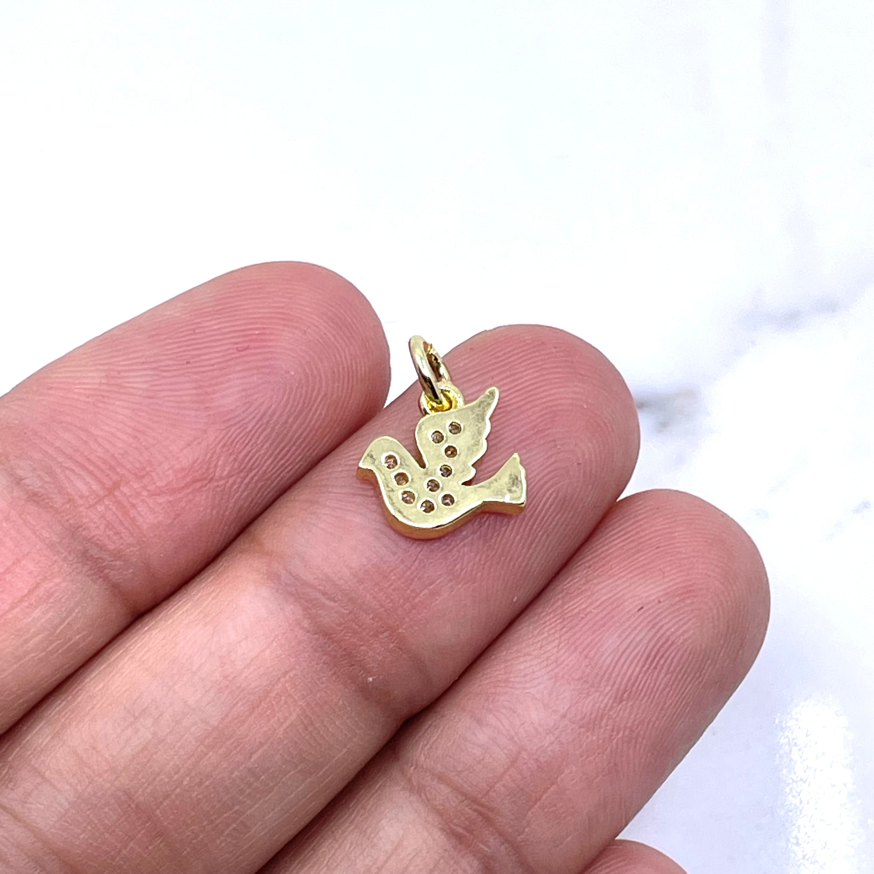 A small gold bird charm with a textured, dotted pattern is held between a person's fingers against a white marble background. (back side of dove charm)