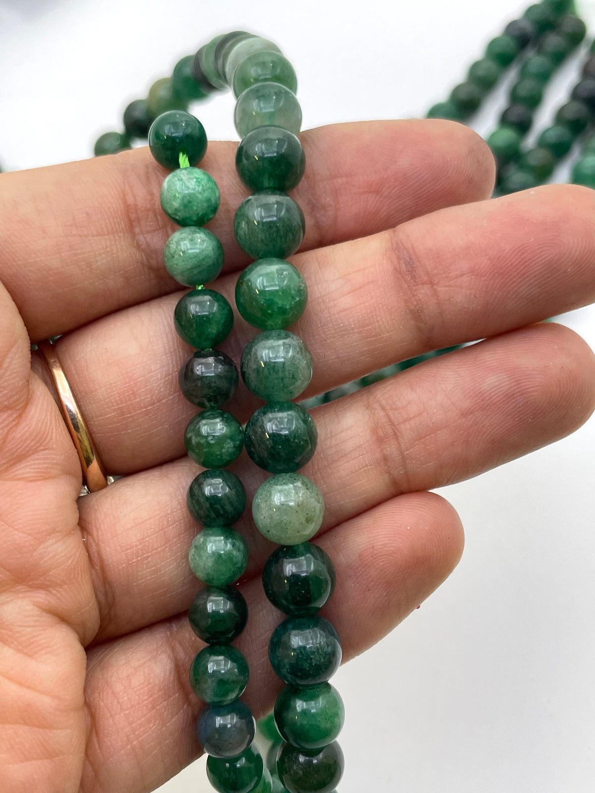 A close-up of a hand holding a strand of green lepidolite gemstone beads, with more matching beads blurred in the background against a white backdrop.