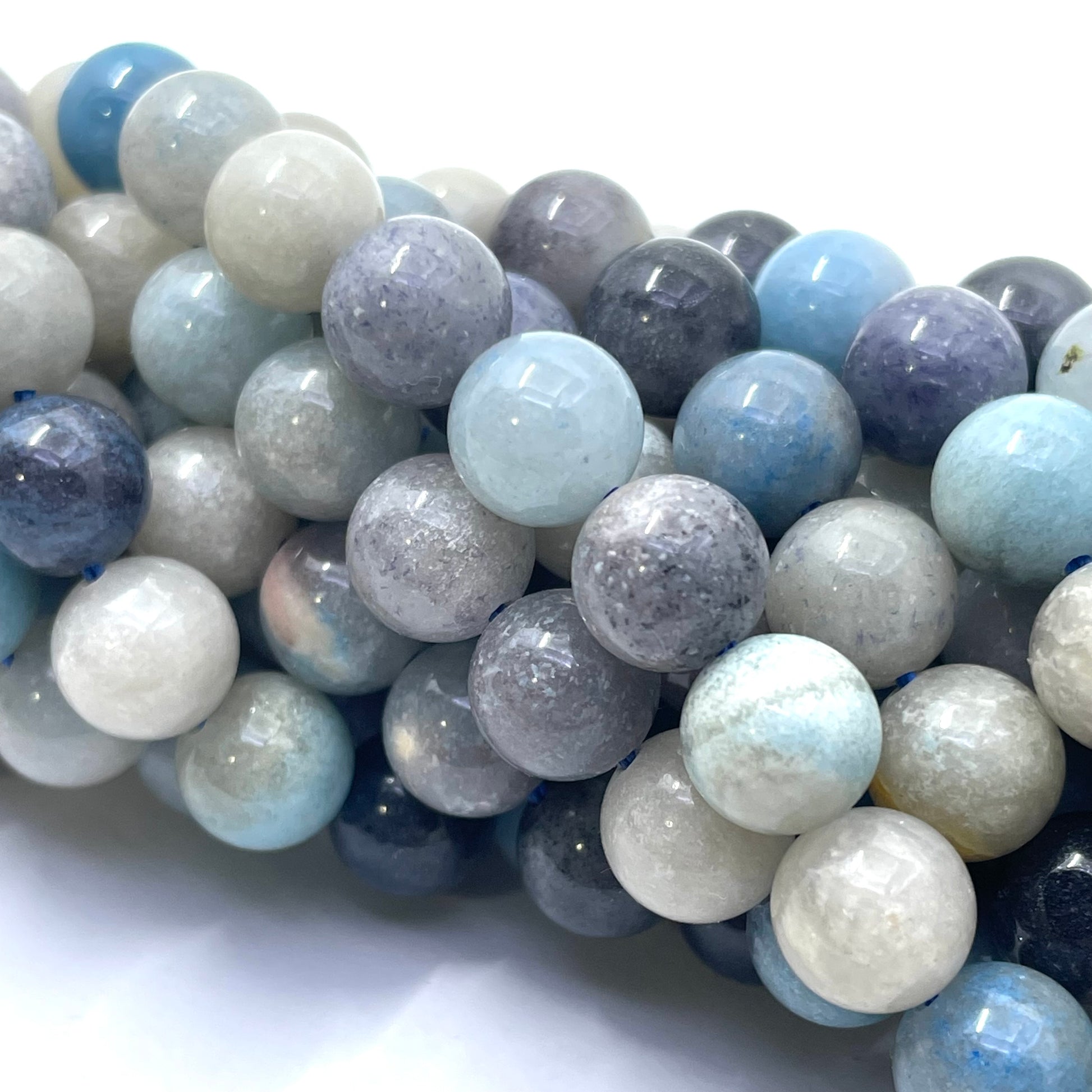 A close-up of multiple strands of round polished trolleite gemstone beads in light blue, white, gray, and lavender-purple tones, arranged diagonally on a white background.