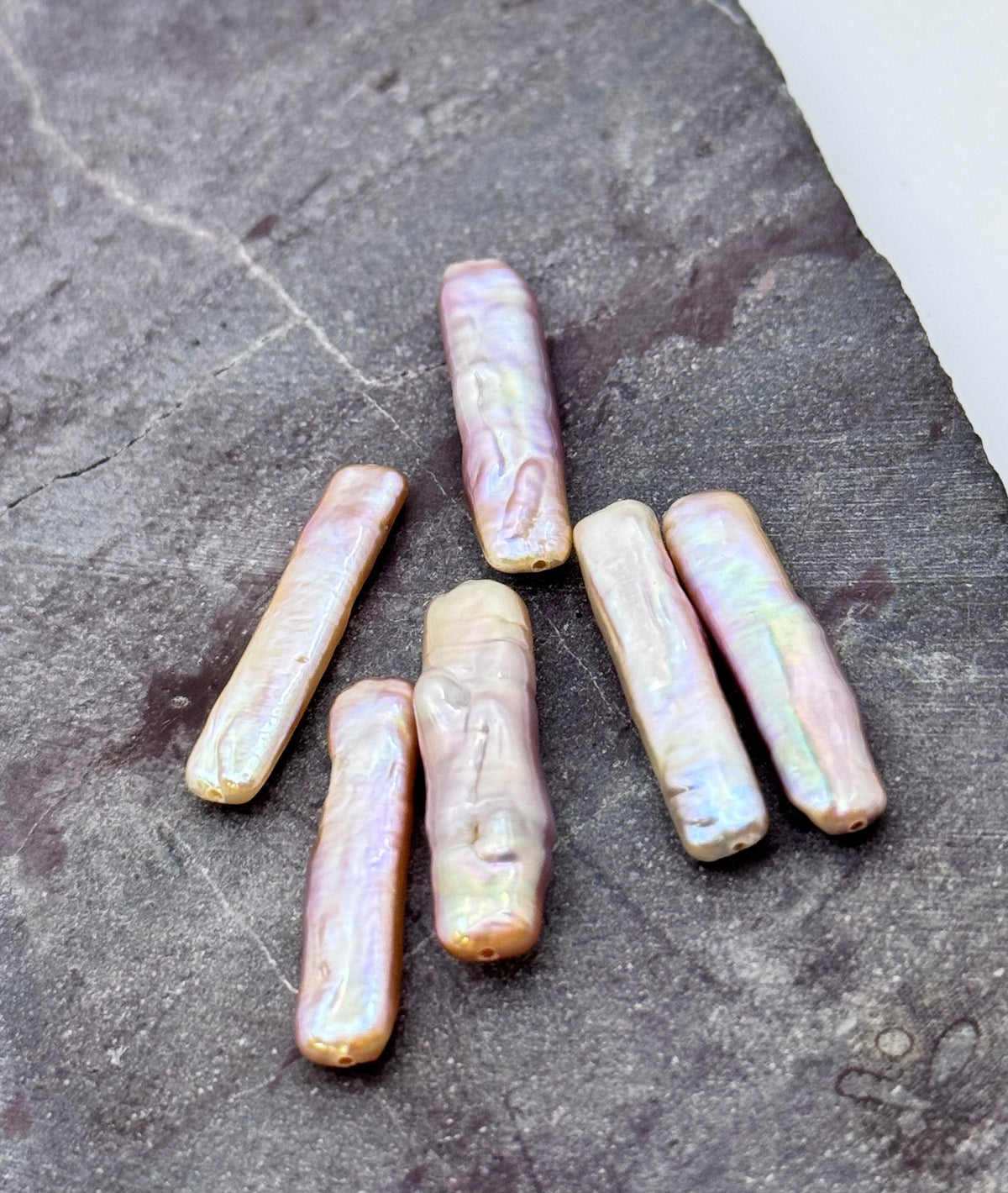 Five baroque freshwater pearl beads with an iridescent lavender-pink sheen, arranged on a dark textured slate surface.