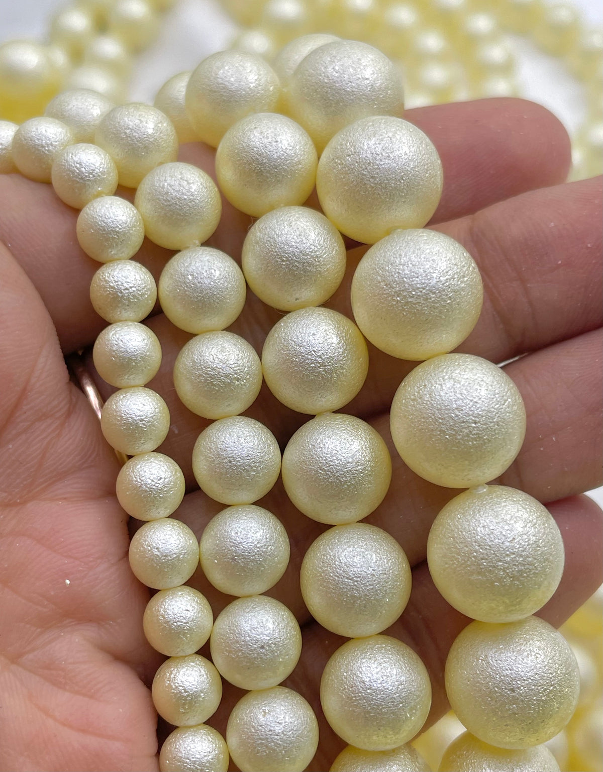 A close-up of a hand holding strands of glossy shell pearl beads in a pale yellow color with a subtle glitter finish.