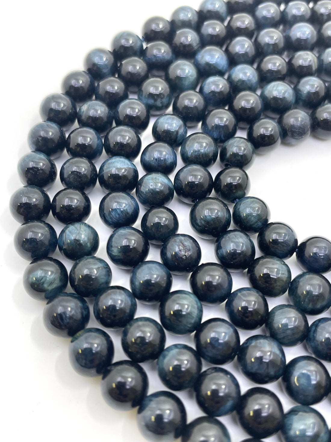 A close-up of glossy dark blue to black round tiger eye beads arranged in strands on a plain white background.