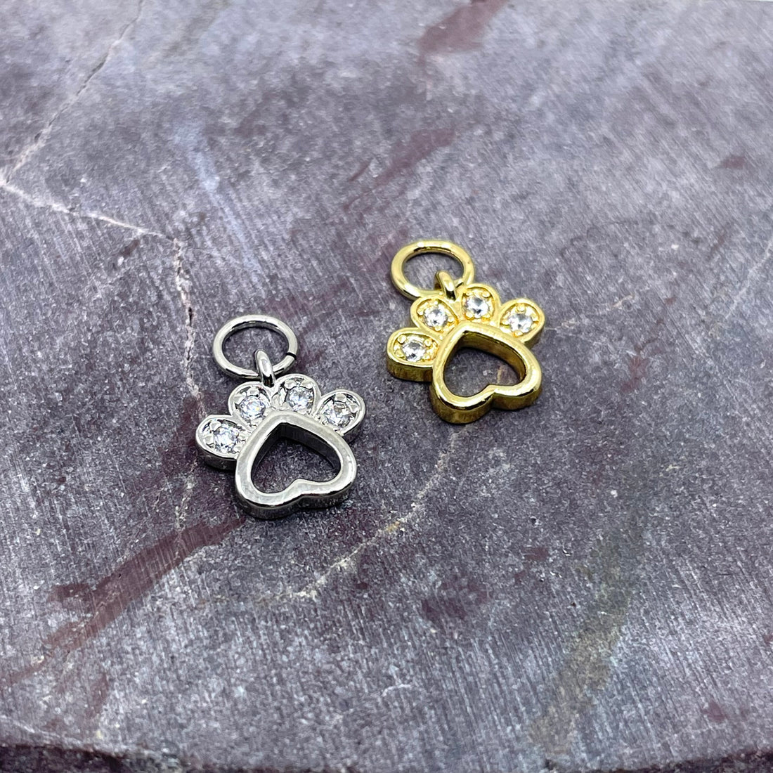 Two small paw-shaped jewelry charms with heart cutouts and clear rhinestone accents, shown on a textured gray stone surface. One charm is silver-toned and the other is gold-toned.