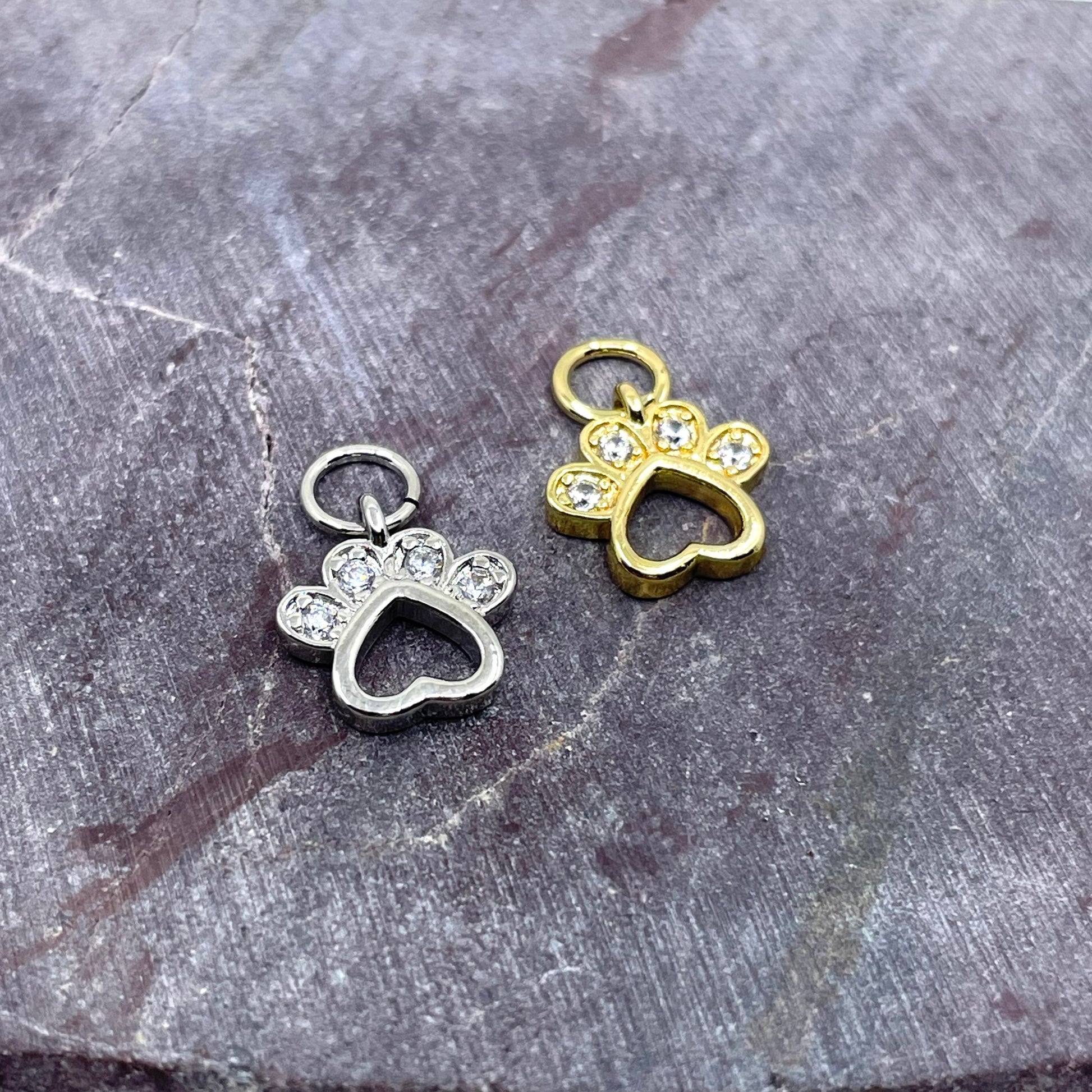 Two small paw-shaped jewelry charms with heart cutouts and clear rhinestone accents, shown on a textured gray stone surface. One charm is silver-toned and the other is gold-toned.