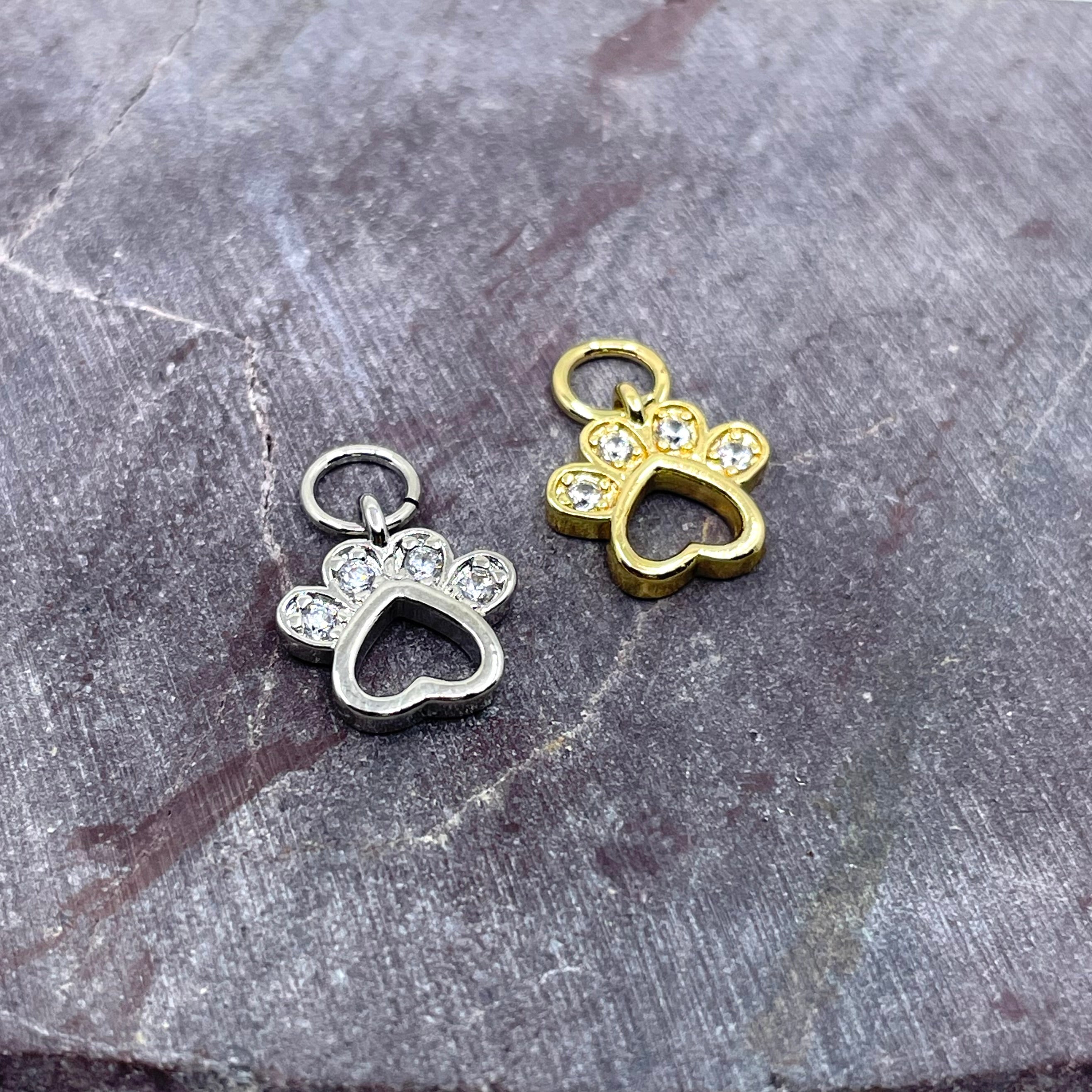 Two small paw-shaped jewelry charms with heart cutouts and clear rhinestone accents, shown on a textured gray stone surface. One charm is silver-toned and the other is gold-toned.