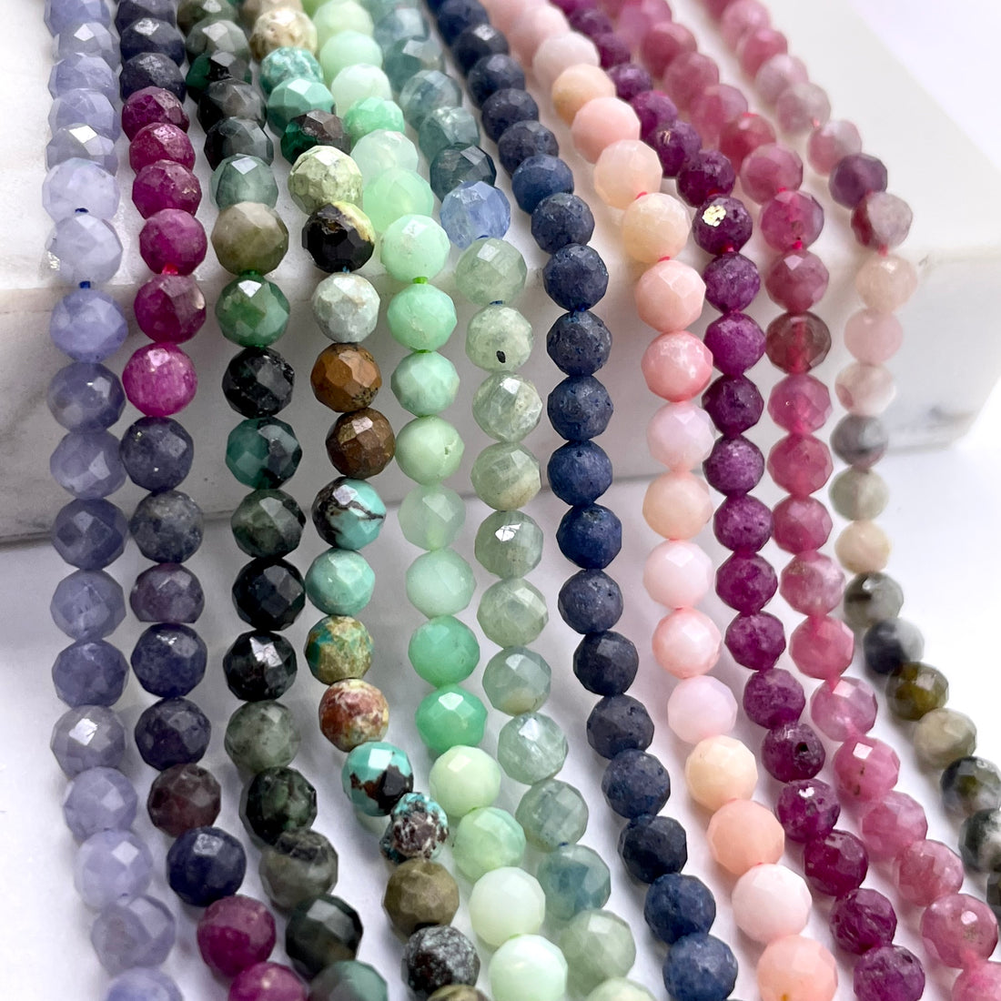 Multiple strands of faceted round gemstone beads in assorted colors, including lavender, green, blue, pink, and purple, arranged vertically on a white surface.