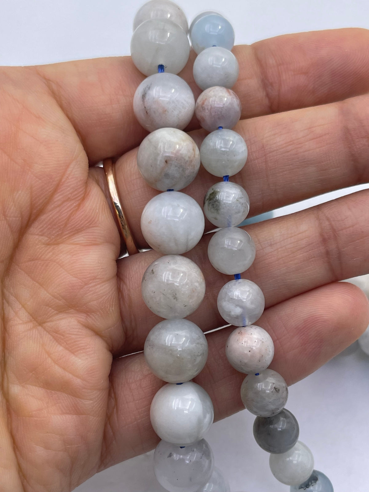 A close-up of a hand holding a strand of round aquamarine white and light gray stone beads, with a rose gold band visible on the ring finger against a plain white background.