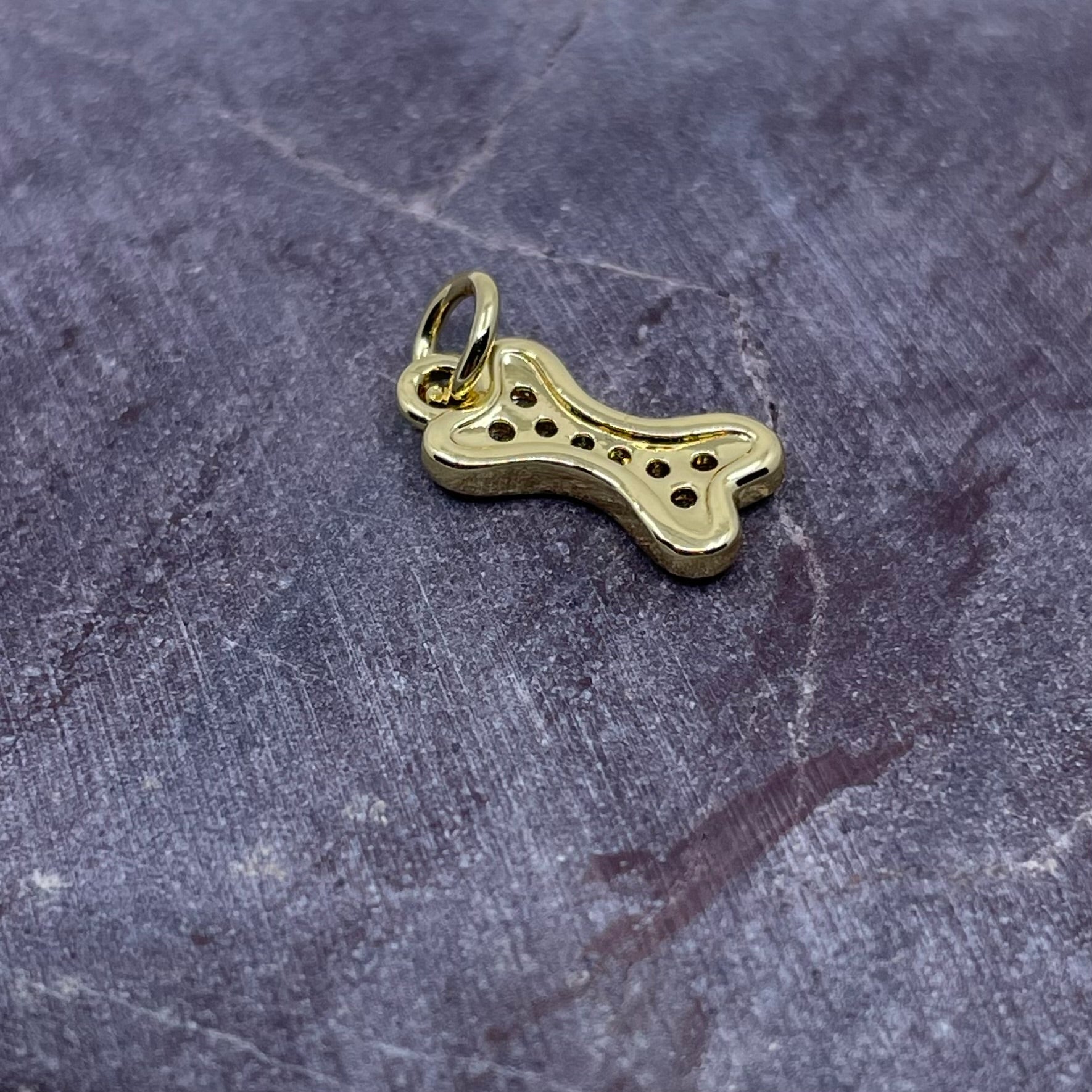 A small gold charm shaped like a dog bone with perforated details, resting on a textured dark gray stone surface.