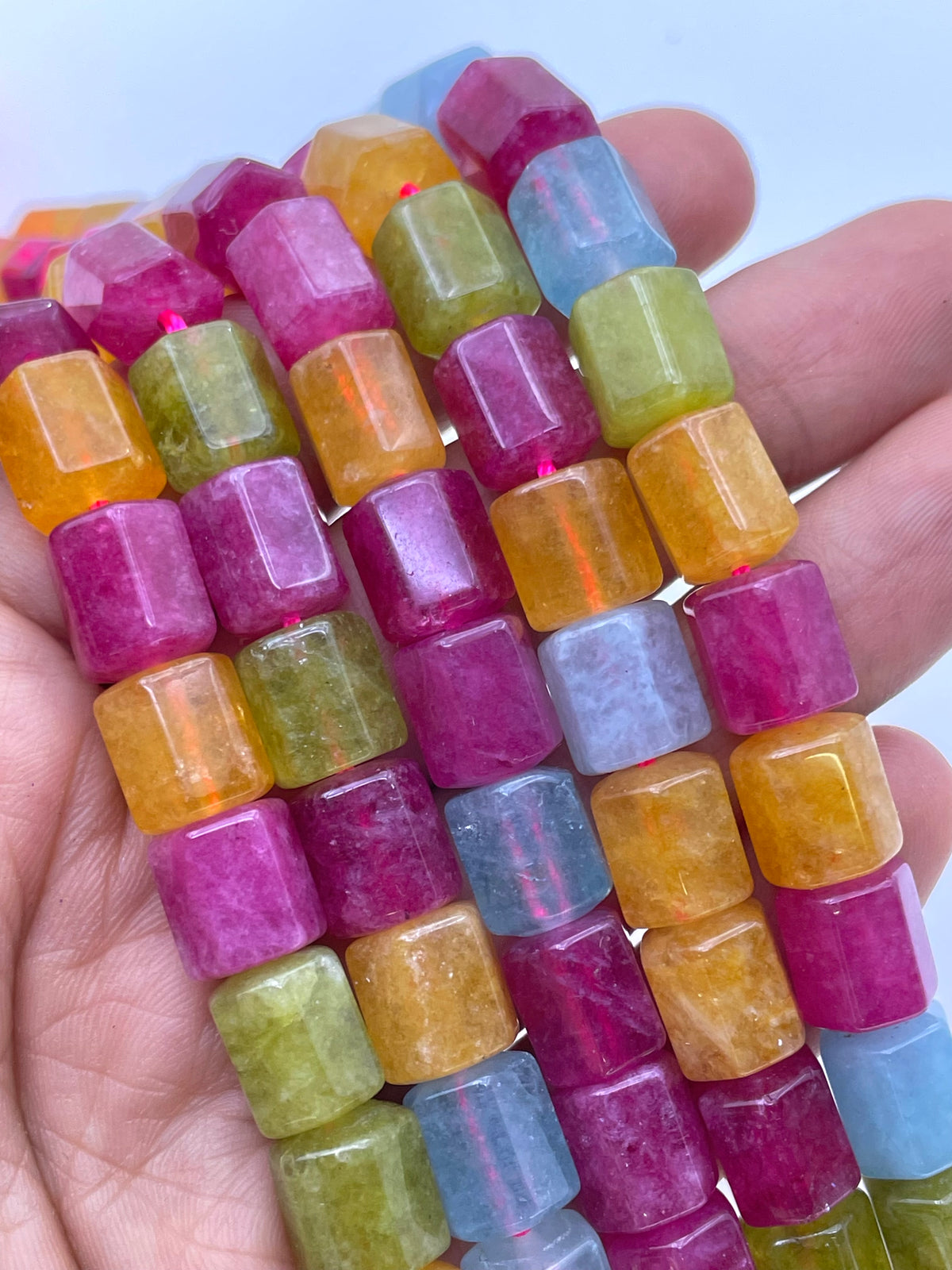 A close-up of a hand holding multiple strands of faceted cylindrical jade gemstone beads in assorted colors including magenta, orange, yellow-green, light blue, and lavender against a pale blue background.