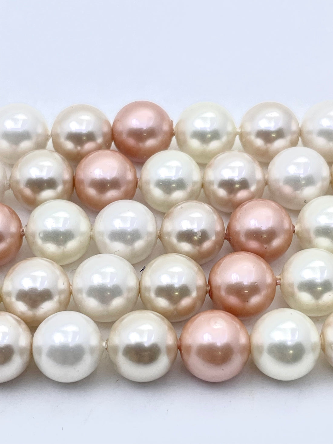Close-up of strands of round shell pearls in white, cream, and soft peach-pink tones on a light gray background.