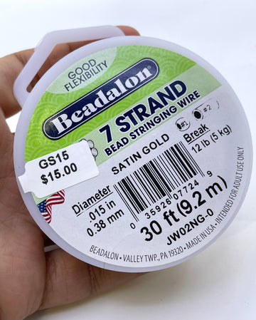 A hand holding a spool of Beadalon 7 Strand Bead Stringing Wire in Satin Gold. The spool is white with a green and blue label showing product details like diameter, length, and price.