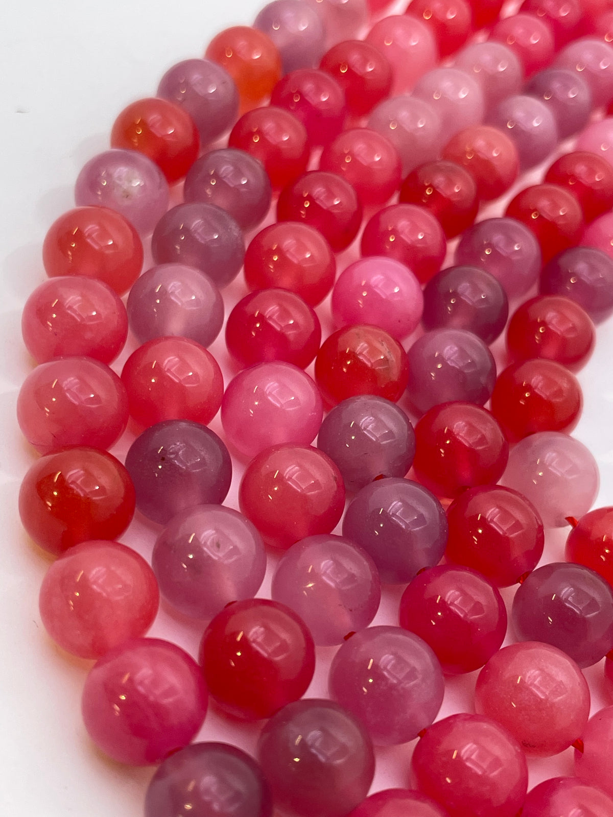 A close-up of glossy round scarlet agate beads in shades of pink, red, and purple, arranged in diagonal rows on a white background.
