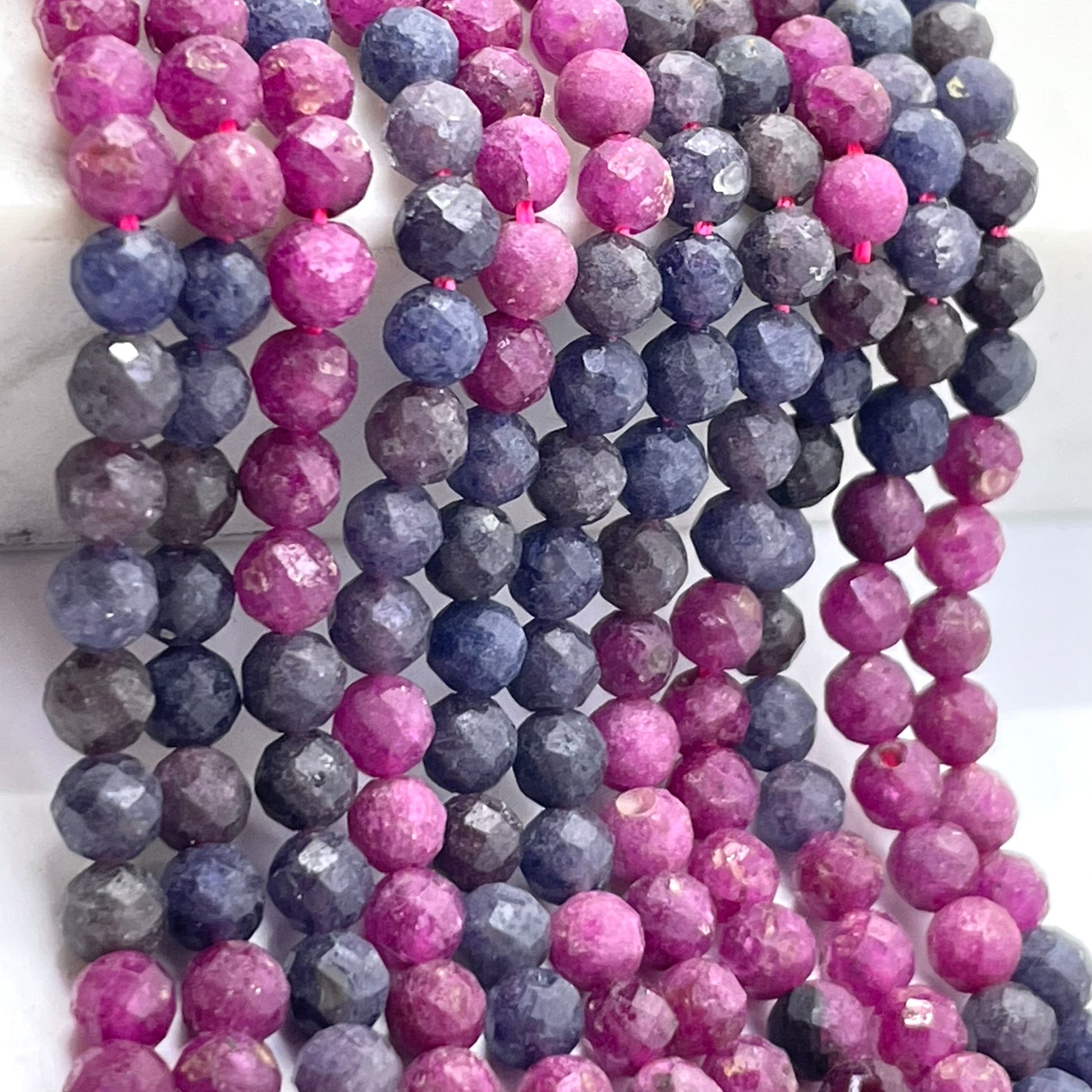 Multiple strands of faceted round ruby sapphire gemstone beads draped vertically, showing pink, purple, and blue-gray tones against a light neutral background.