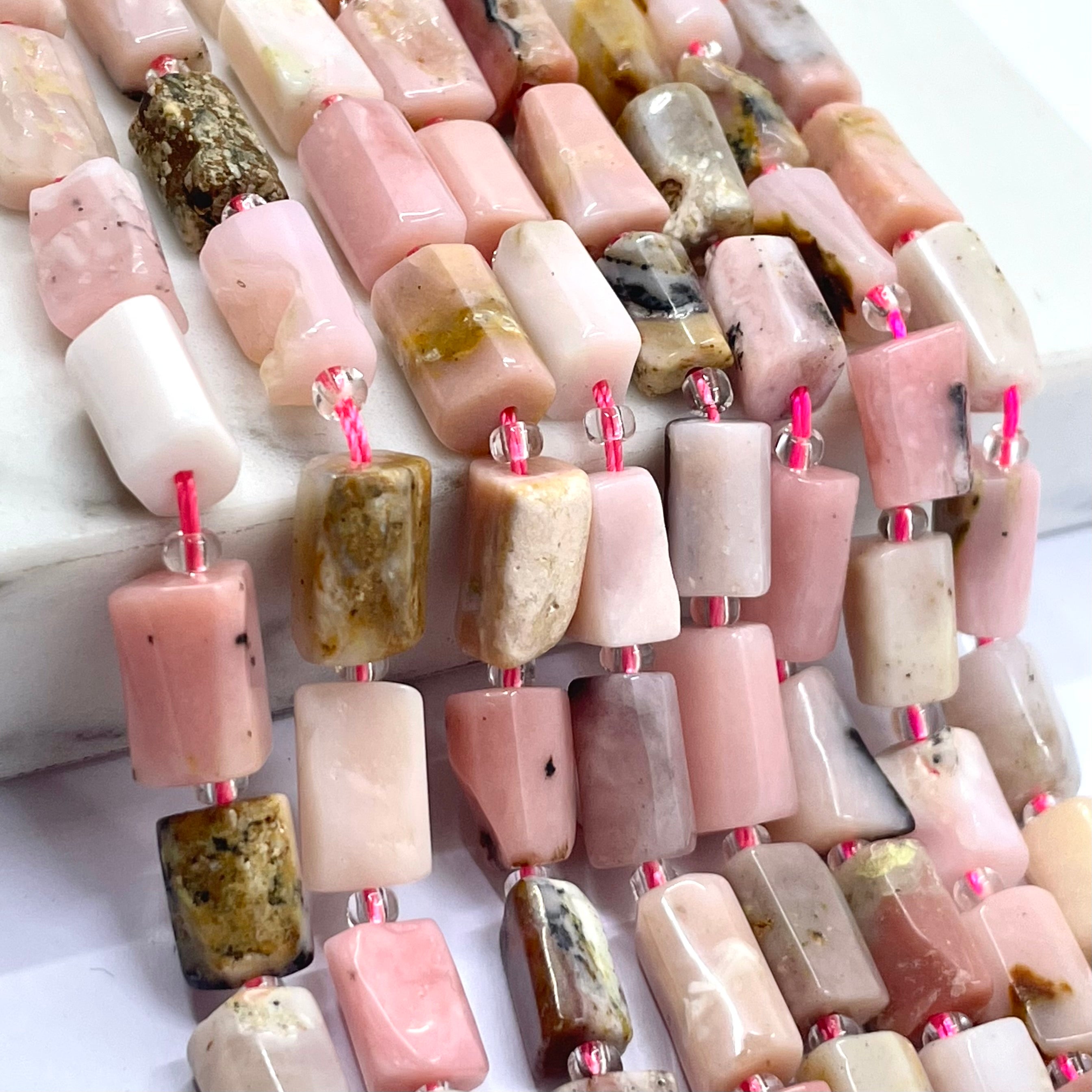 Close-up of multiple strands of rectangular polished pink opal stone beads in soft pink, blush, cream, and earthy brown tones, strung with small clear beads and bright pink cord.