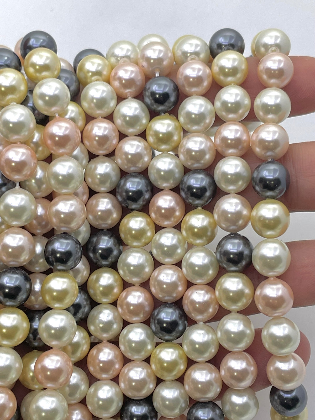 Multiple strands of shell pearl beads in assorted colors including cream, peach, pink, gray, and black, held in a hand against a plain light background.