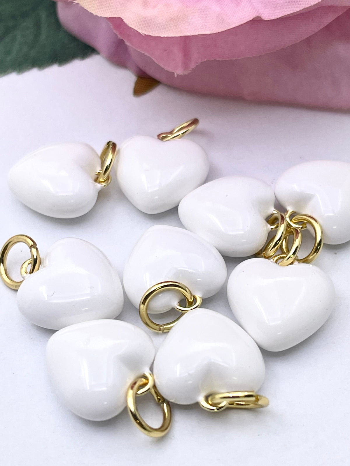 White glossy heart-shaped charms with gold-tone jump rings, arranged on a light surface with a soft pink flower in the background.
