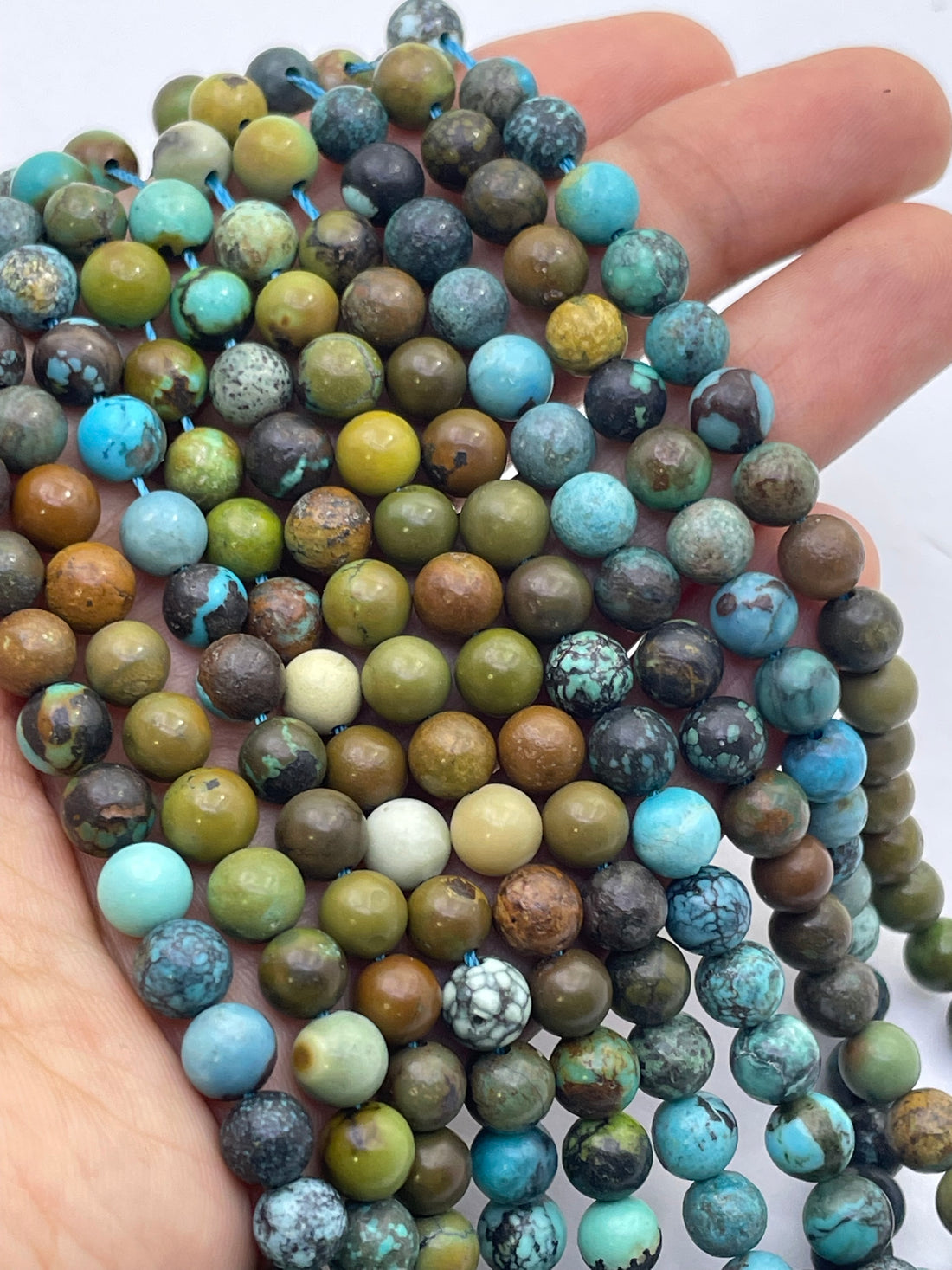 A close-up of a hand holding multiple strands of round turquoise stone beads in various earthy green, brown, blue, and turquoise colors.